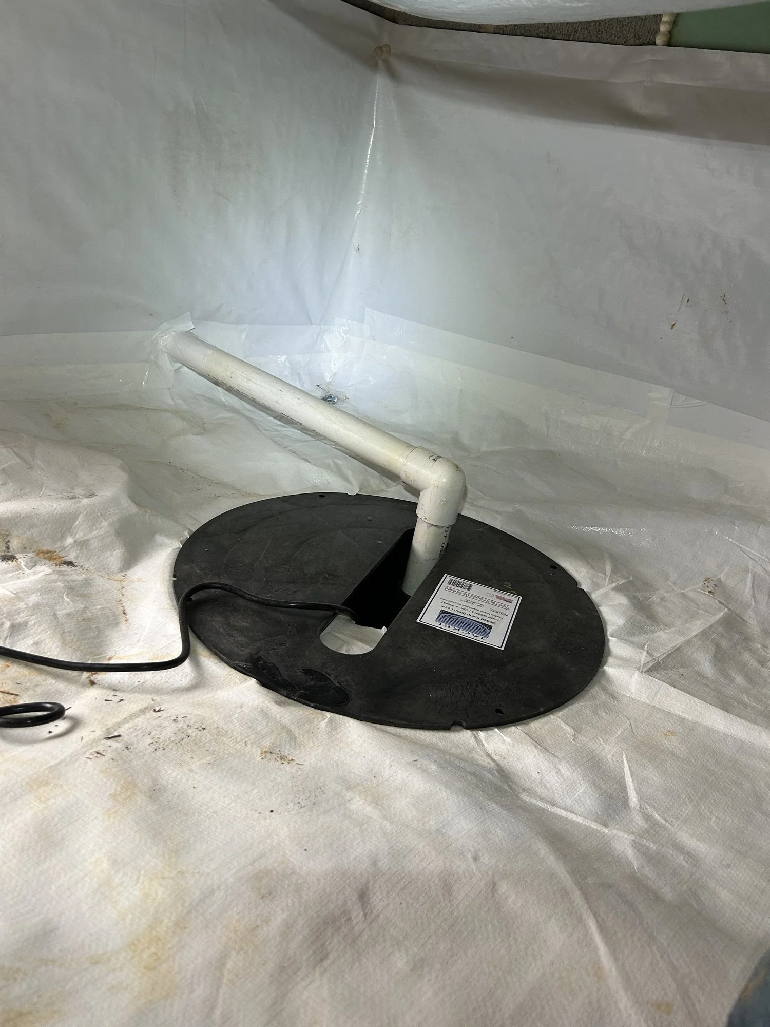 A black plastic base with a white PVC pipe attached, situated on a stained white surface that appears to be fabric or protective covering, with some black electrical cords nearby.