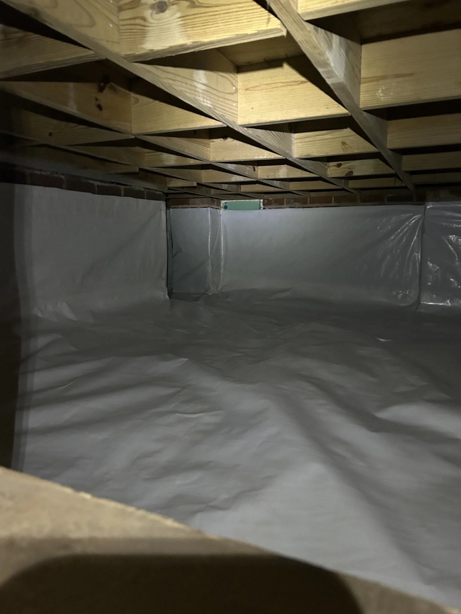 Finish of a basement with a wooden floor above, insulation paper on the floor, and brick walls.