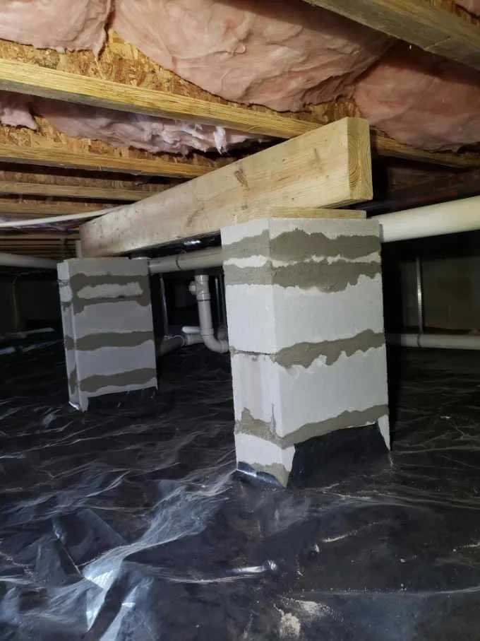 View of a building's crawl space or basement area with concrete block support columns, wooden beams, pink insulation material, and plumbing pipes, all located underneath the floor.