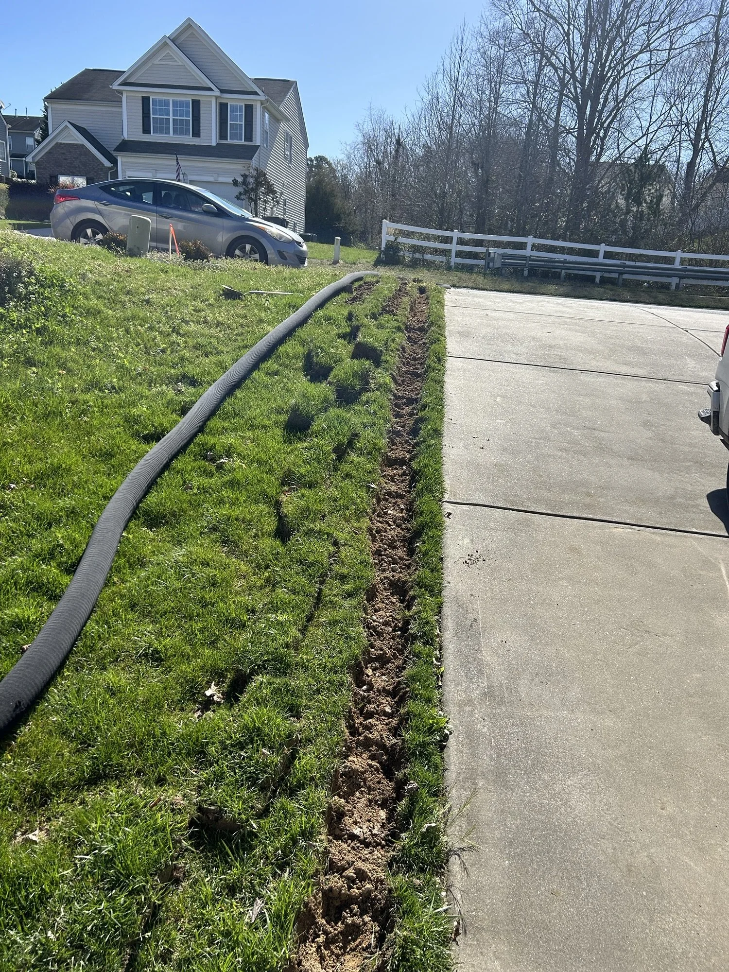 A trench dug along the edge of a concrete driveway in front of a white house with a gray roof. A black flexible pipe runs along the trench. There is a grassy lawn beside the trench, and a house with an American flag is in the background. The sky is clear and sunny.