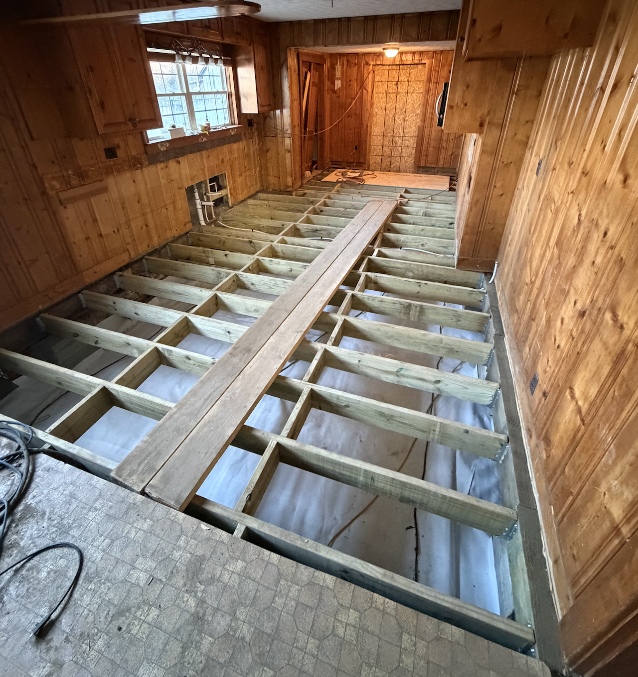 Room with wooden walls and ceiling under renovation, with flooring removed to expose floor joists and subfloor, and a central wooden plank for support.