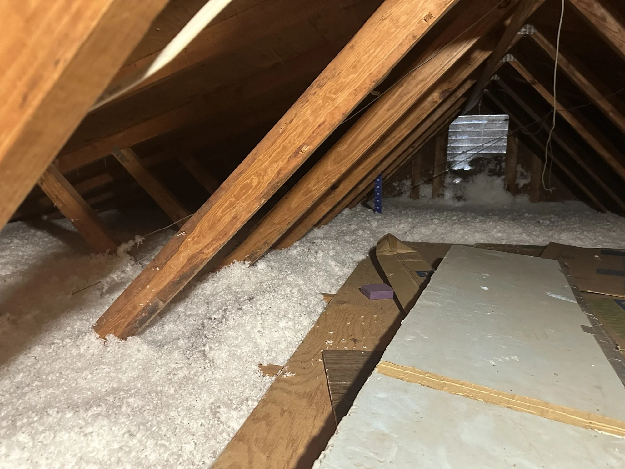 Attic with wooden roof trusses, insulation material, some scattered drywall sheets, and exposed wiring.