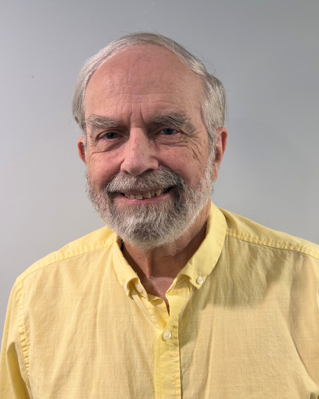 Close-up of an elderly man with gray hair and beard, smiling, wearing a yellow button-down shirt against a plain background.