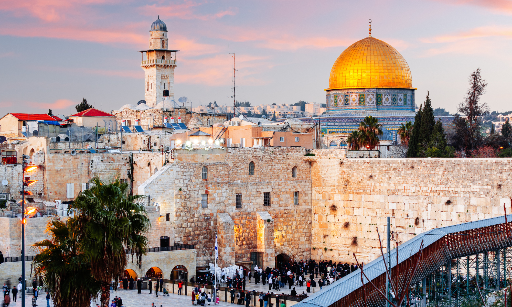 A cityscape of Jerusalem featuring the Western Wall, with the Dome of the Rock and its golden dome in the background during sunset. There are people gathered near the wall and trees in the scene.