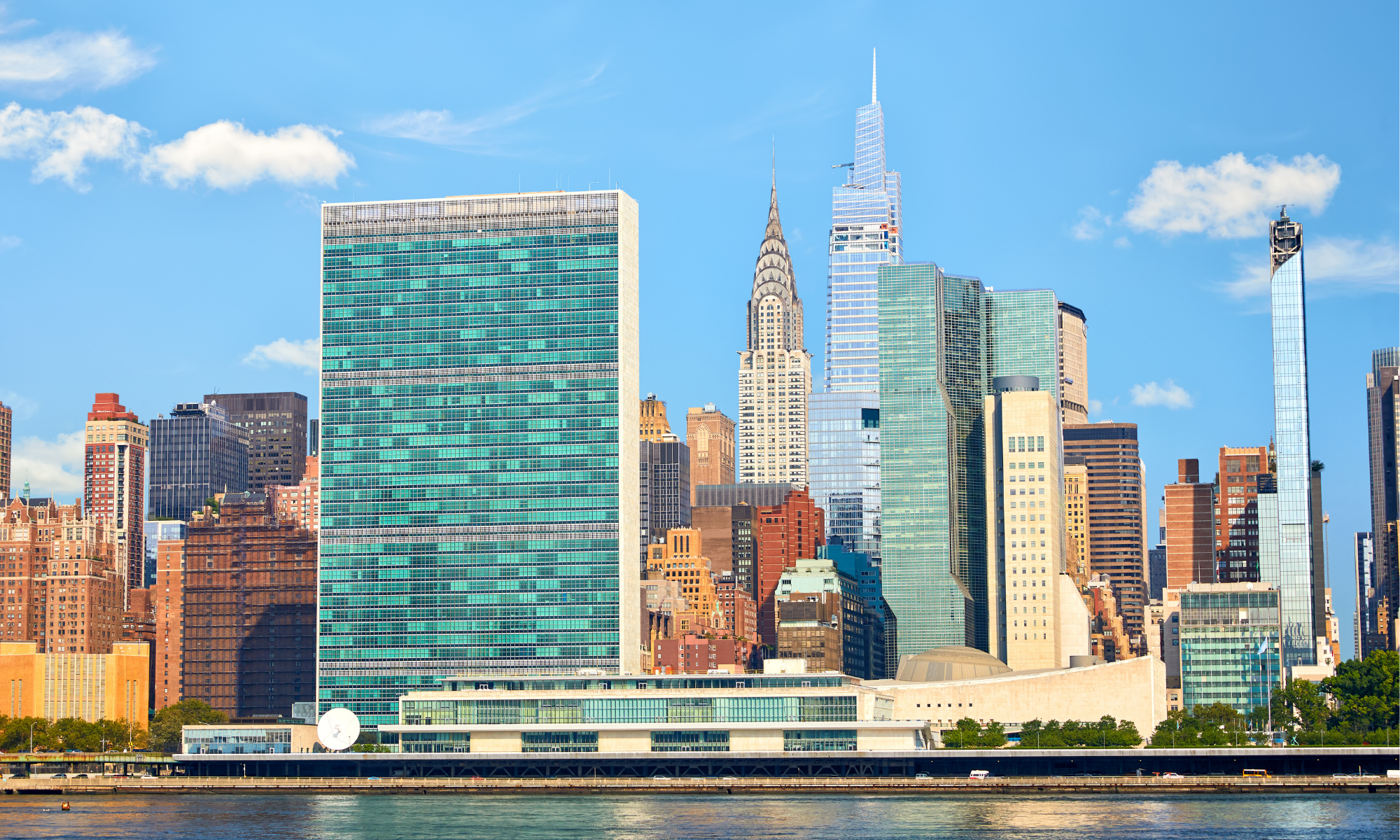 Skyline of New York City with tall skyscrapers including the Chrysler Building and One Vanderbilt, viewed from across the river under a clear blue sky with a few clouds.