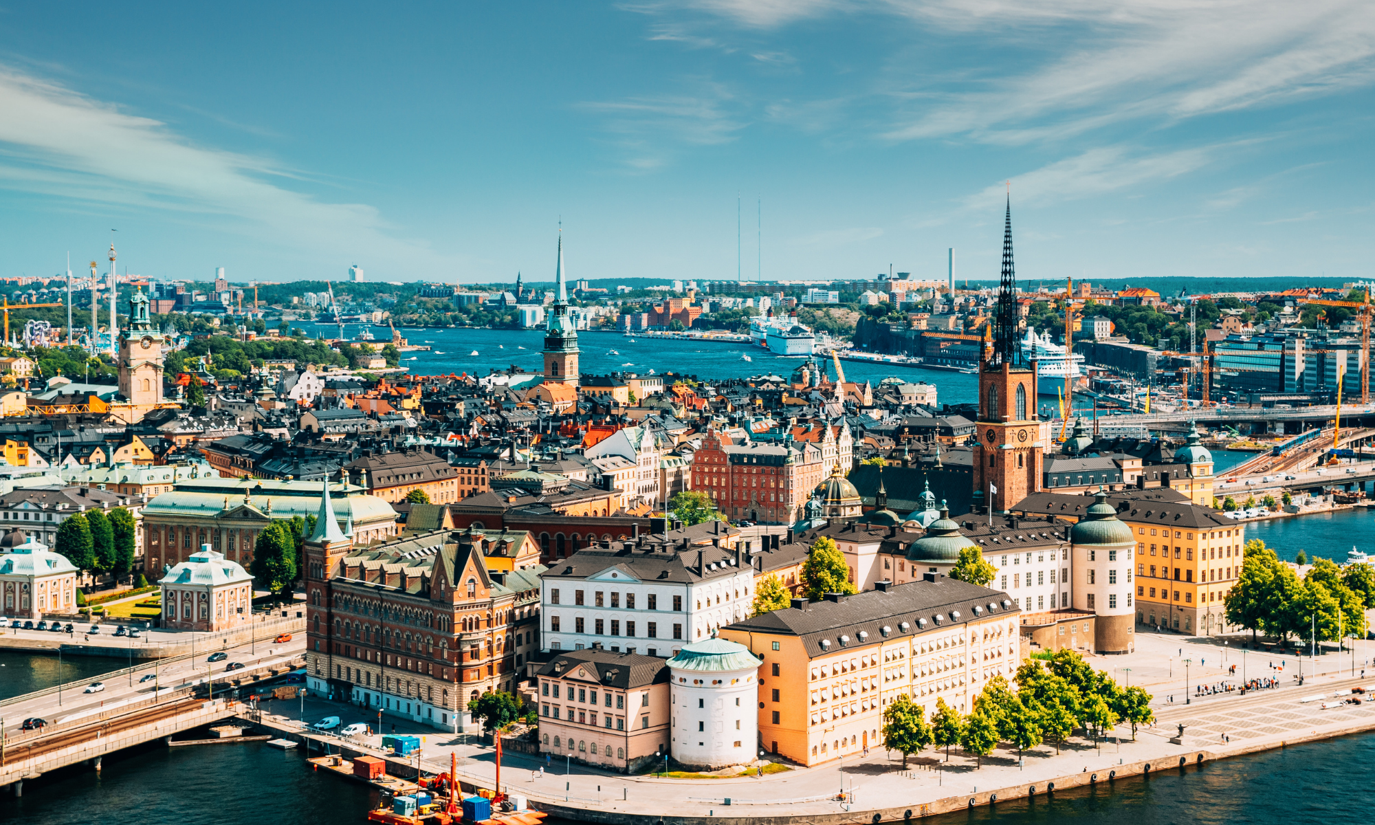 A panoramic cityscape of Stockholm, Sweden, showing historic and modern buildings, churches with steeples, bridges, and boats on the water under a partly cloudy sky.