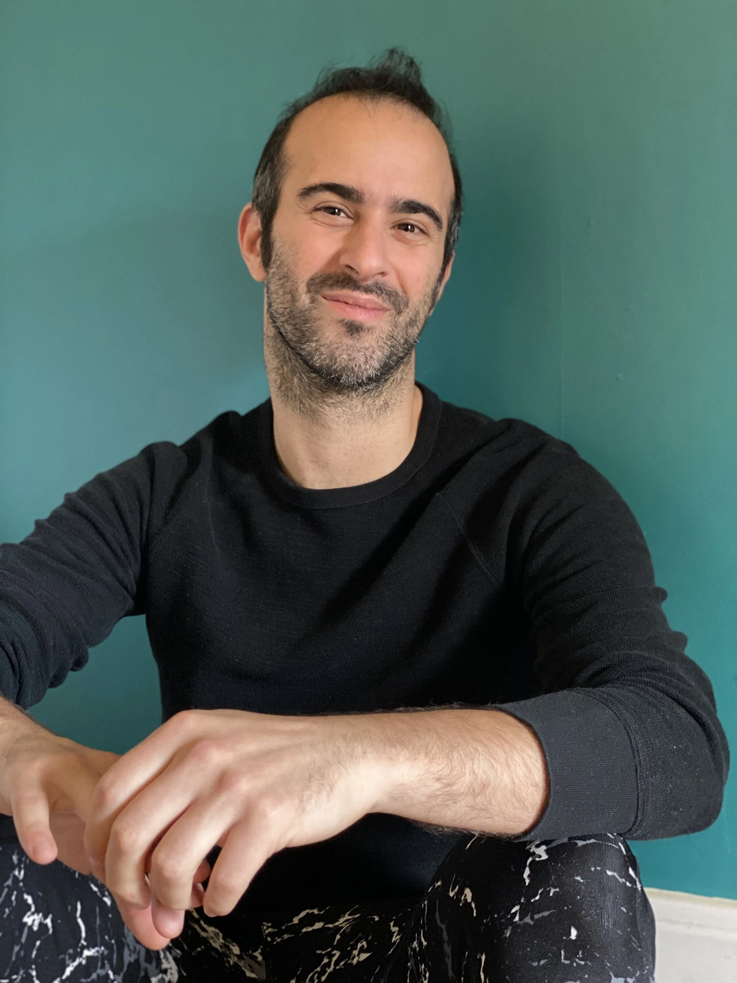 Lolo sits in front of a teal wall in a black shift and black and white leggings, smiling