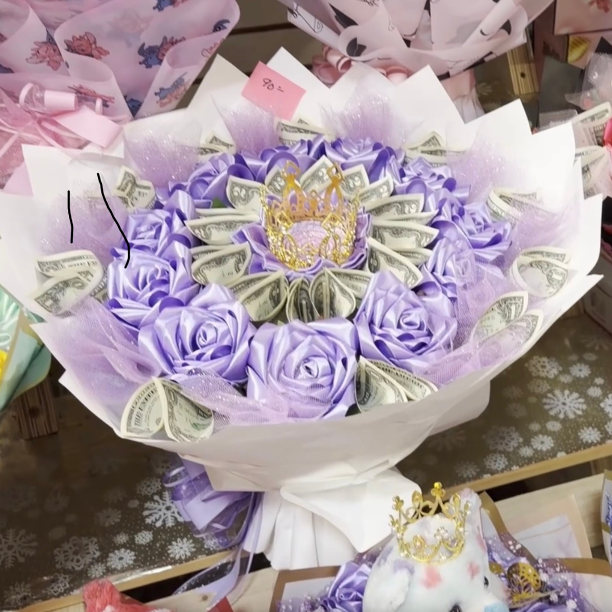 A decorative bouquet made of purple ribbon roses, folded dollar bills, and a gold crown ornament, wrapped in white and purple paper, on a table with snowflake patterns.
