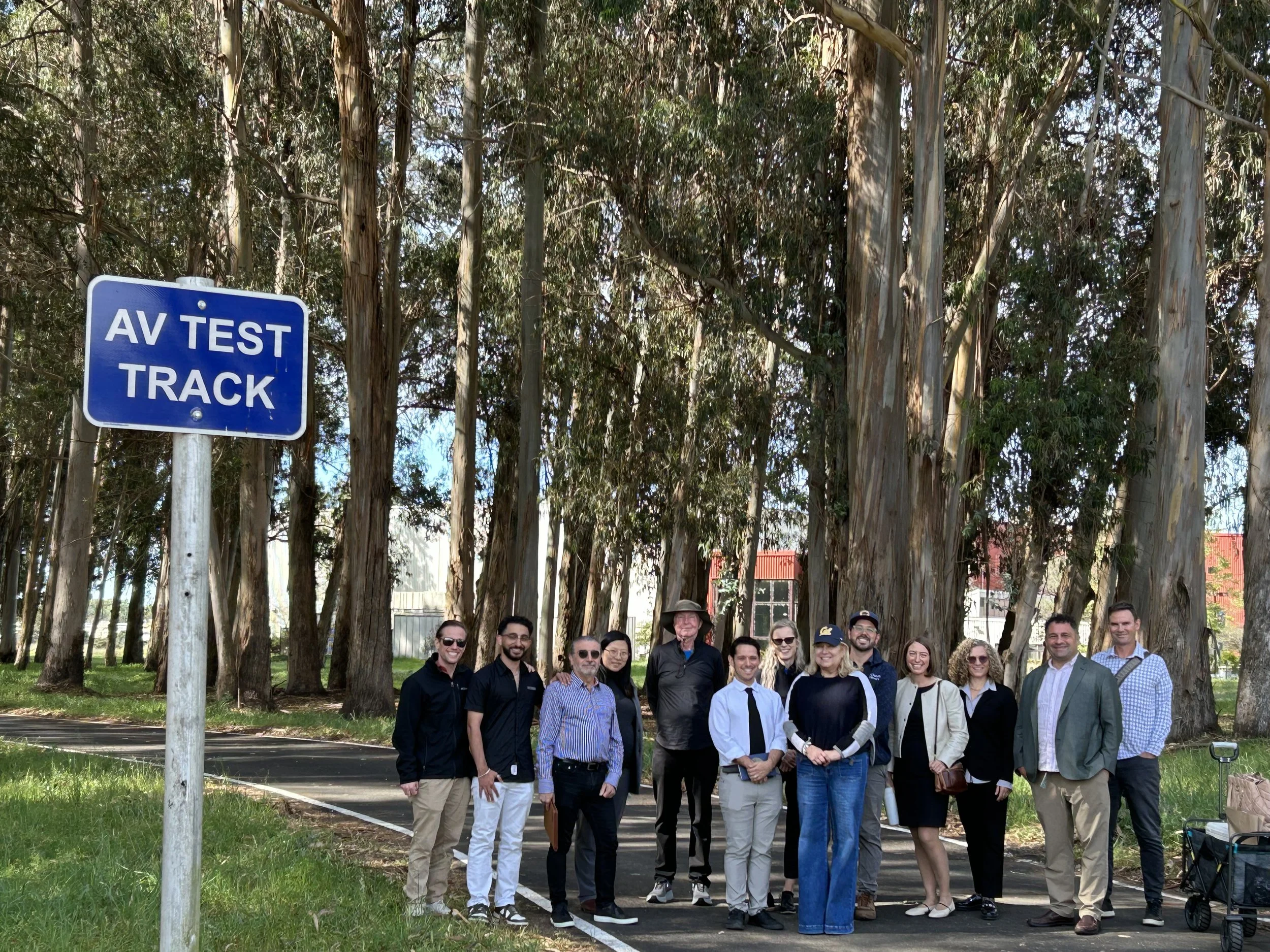 Group photo of Seval Oz and attendees in front of trees with a blue "AV Test Track" sign