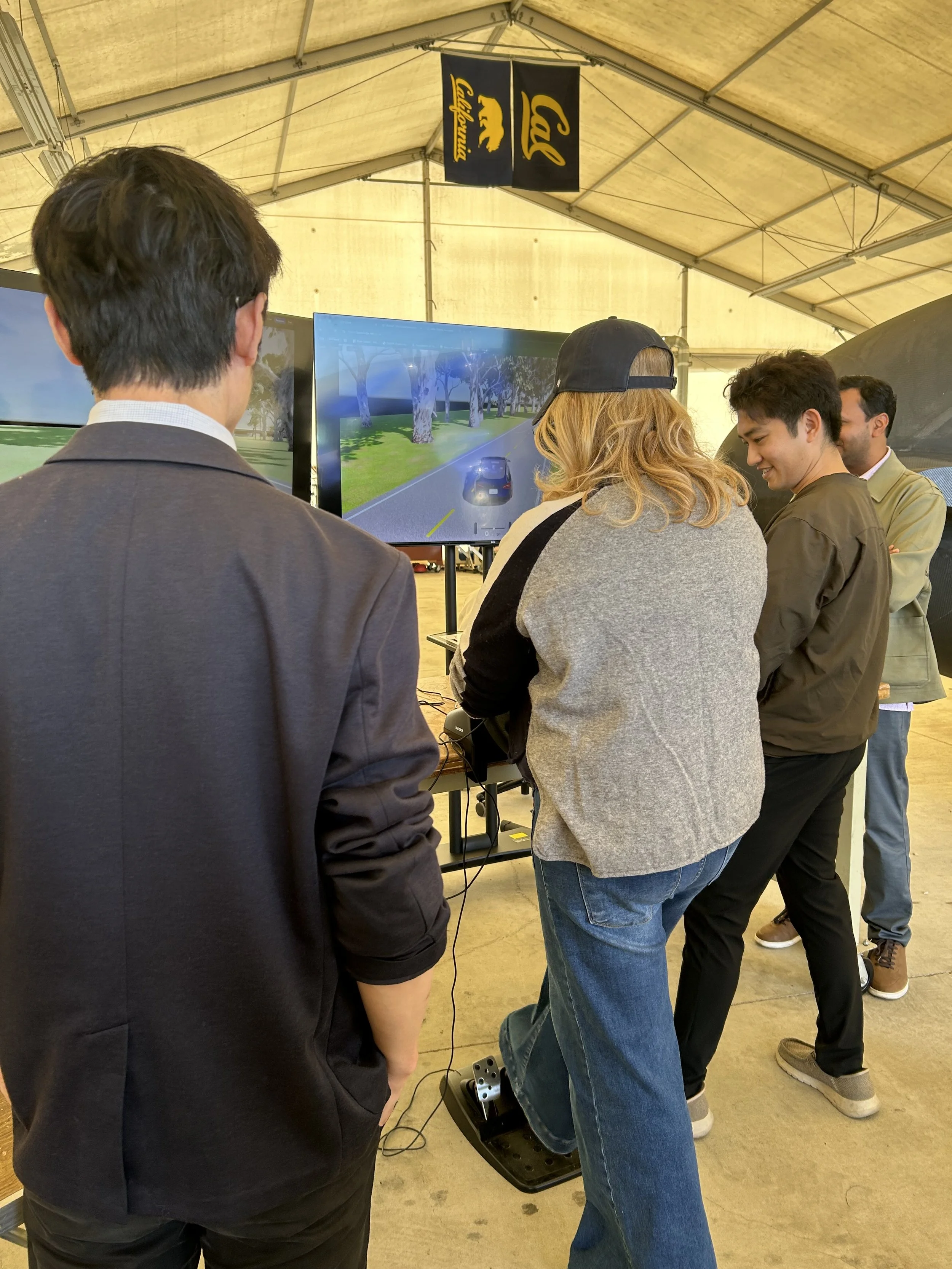 A group of people looking at a screen while one uses a driving simulator. Their backs are to the camera.