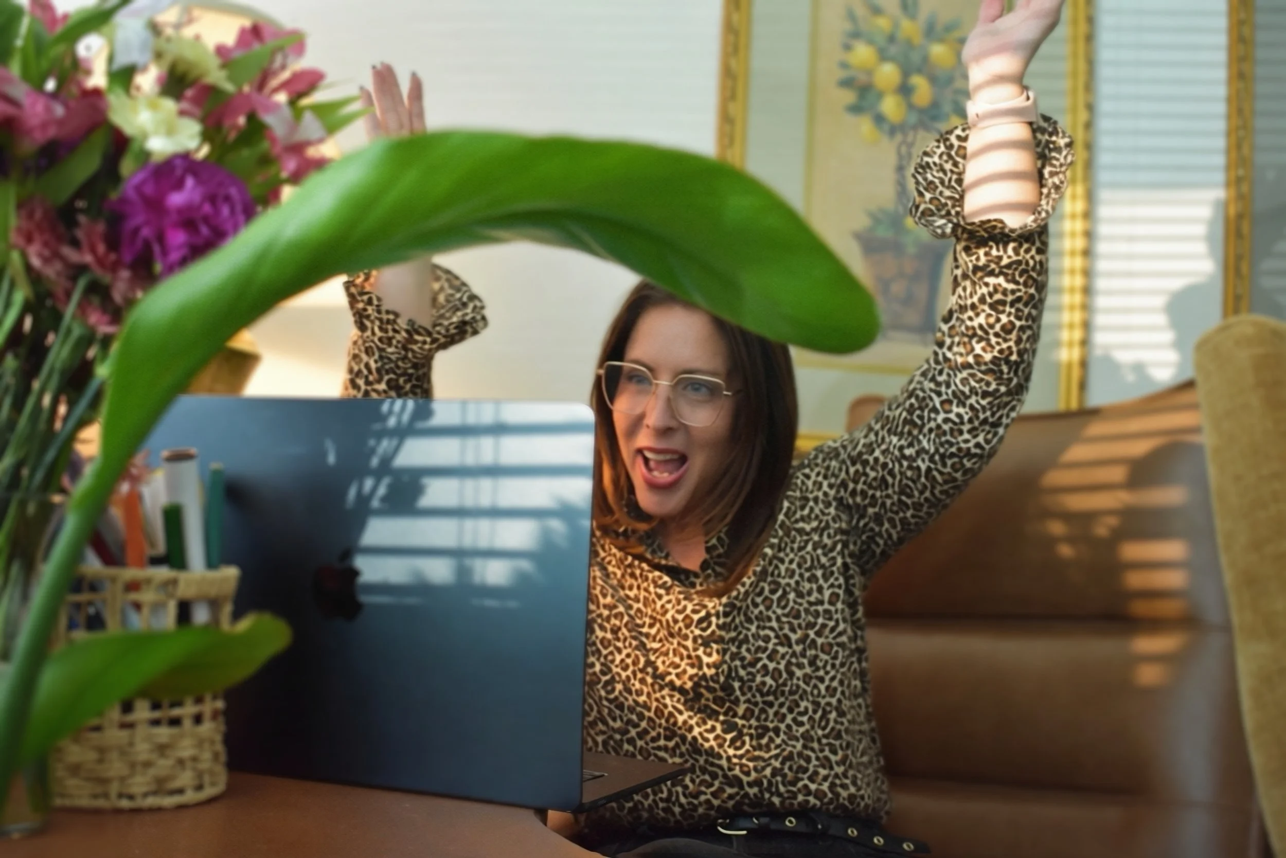 Woman with long brown hair and glasses, wearing a leopard print top, sitting at a desk during a video call or virtual meeting, with a large green plant and a floral bouquet in the foreground.