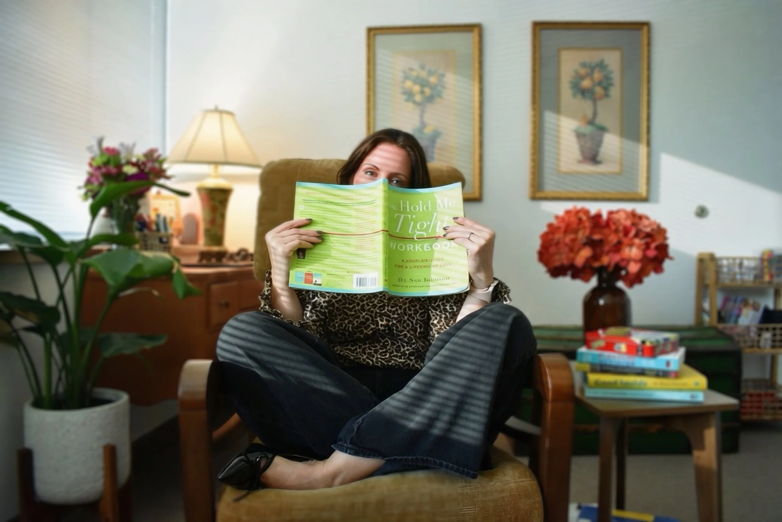 Woman sitting in a cozy living room, reading a green workbook titled 'Hold Me Tight' with a patterned black and white outfit, surrounded by plants, flowers, and framed artwork on the wall.