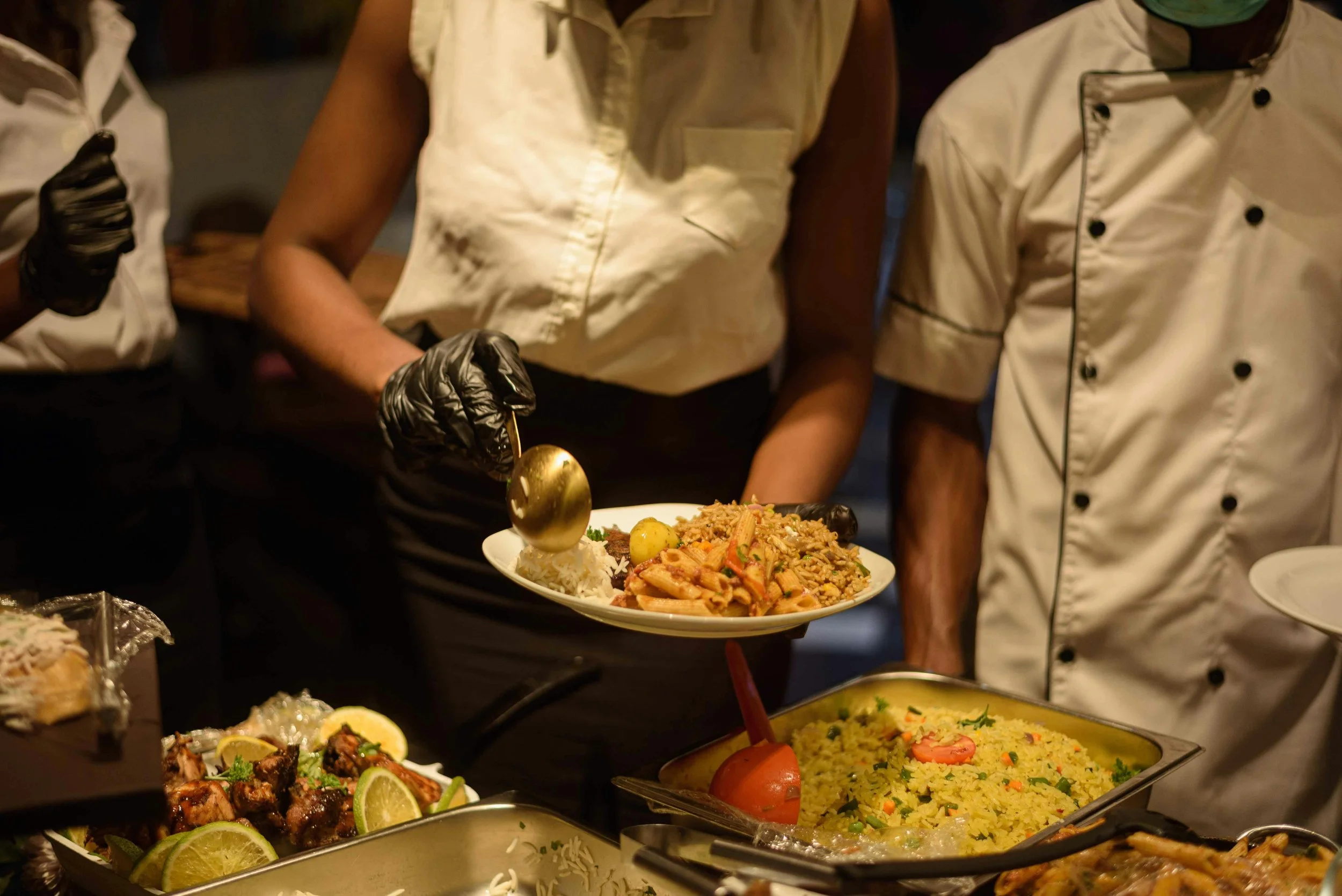 People serving themselves food at a buffet table with dishes including rice, pasta, and meats, with someone holding a plate of rice and pasta.