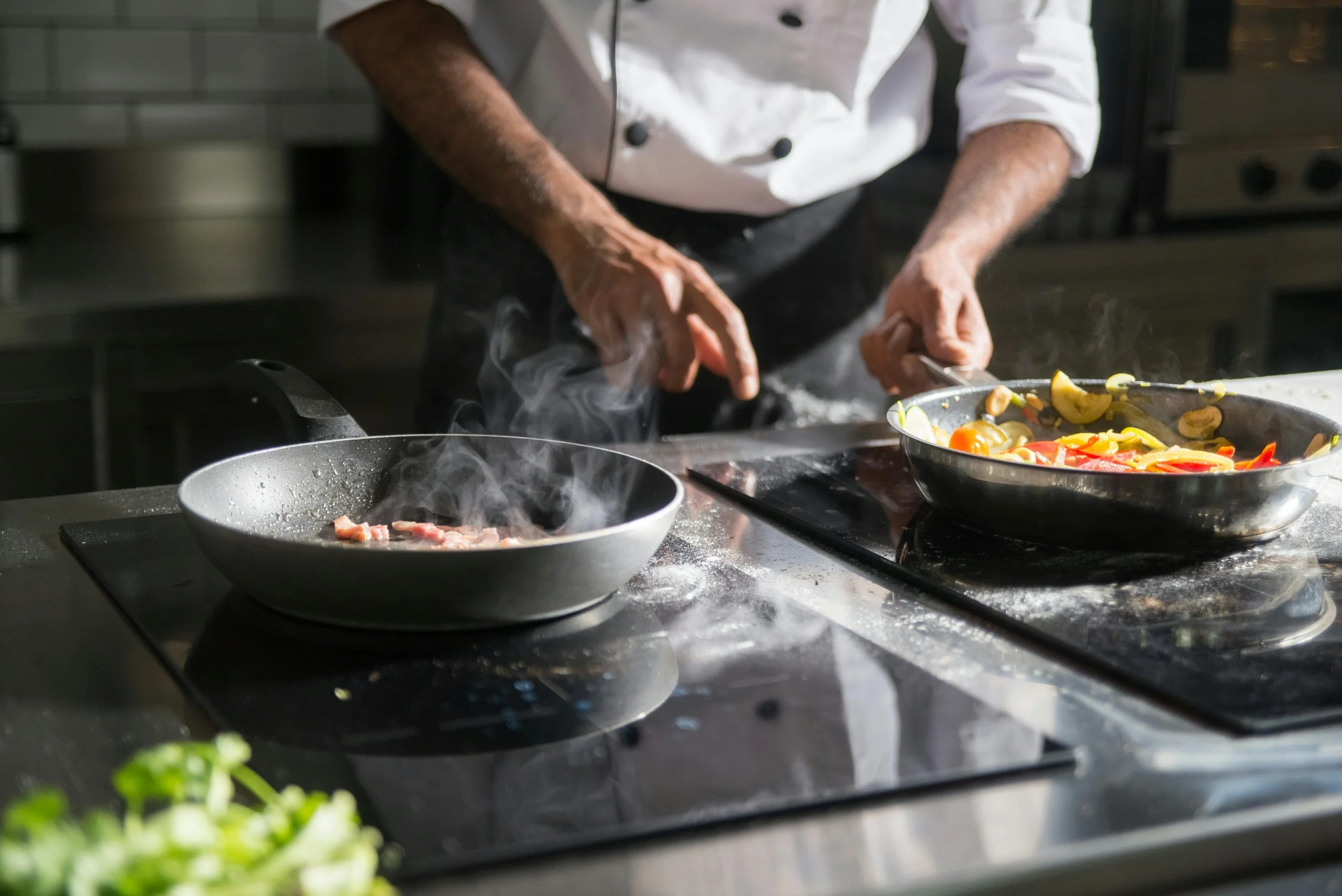 A chef in a white uniform is cooking on a stovetop, preparing a dish with sliced vegetables in a frying pan and some meat in another pan nearby.