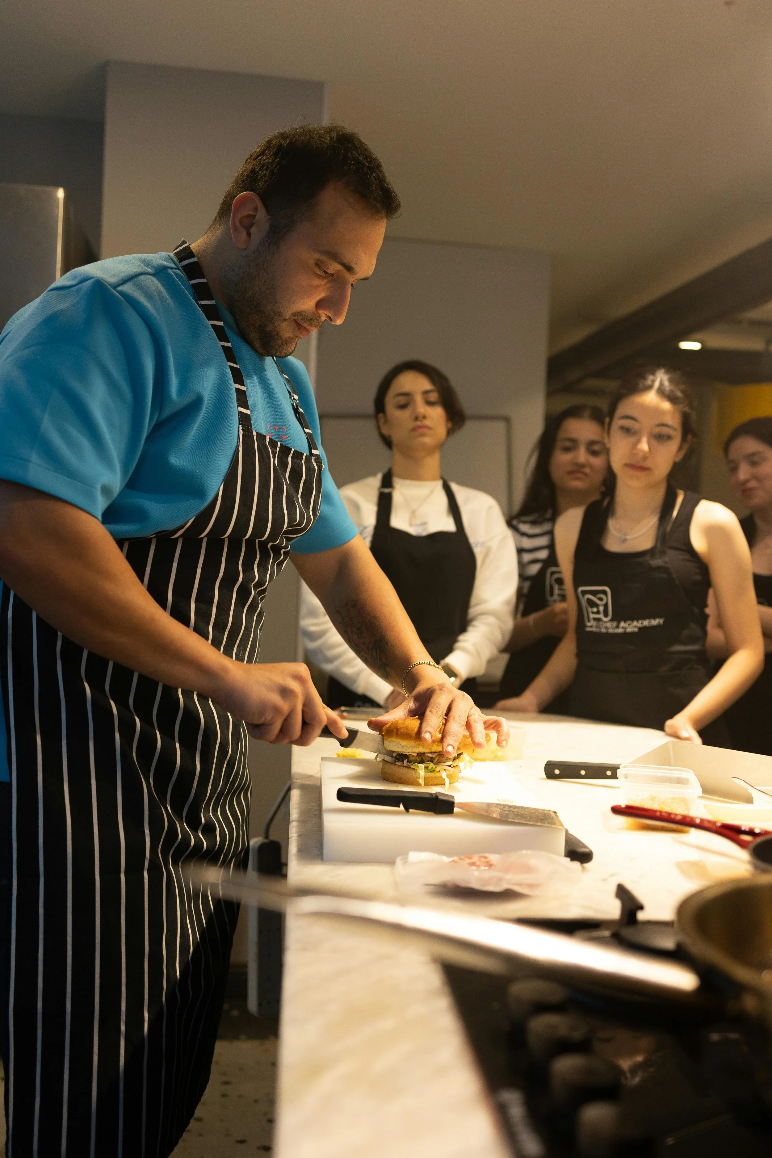 A man with a beard and short dark hair, wearing a blue shirt and a black-and-white striped apron, slices a sandwich on a cutting board at a kitchen counter. Several women, some wearing aprons, watch attentively behind him in a well-lit kitchen.