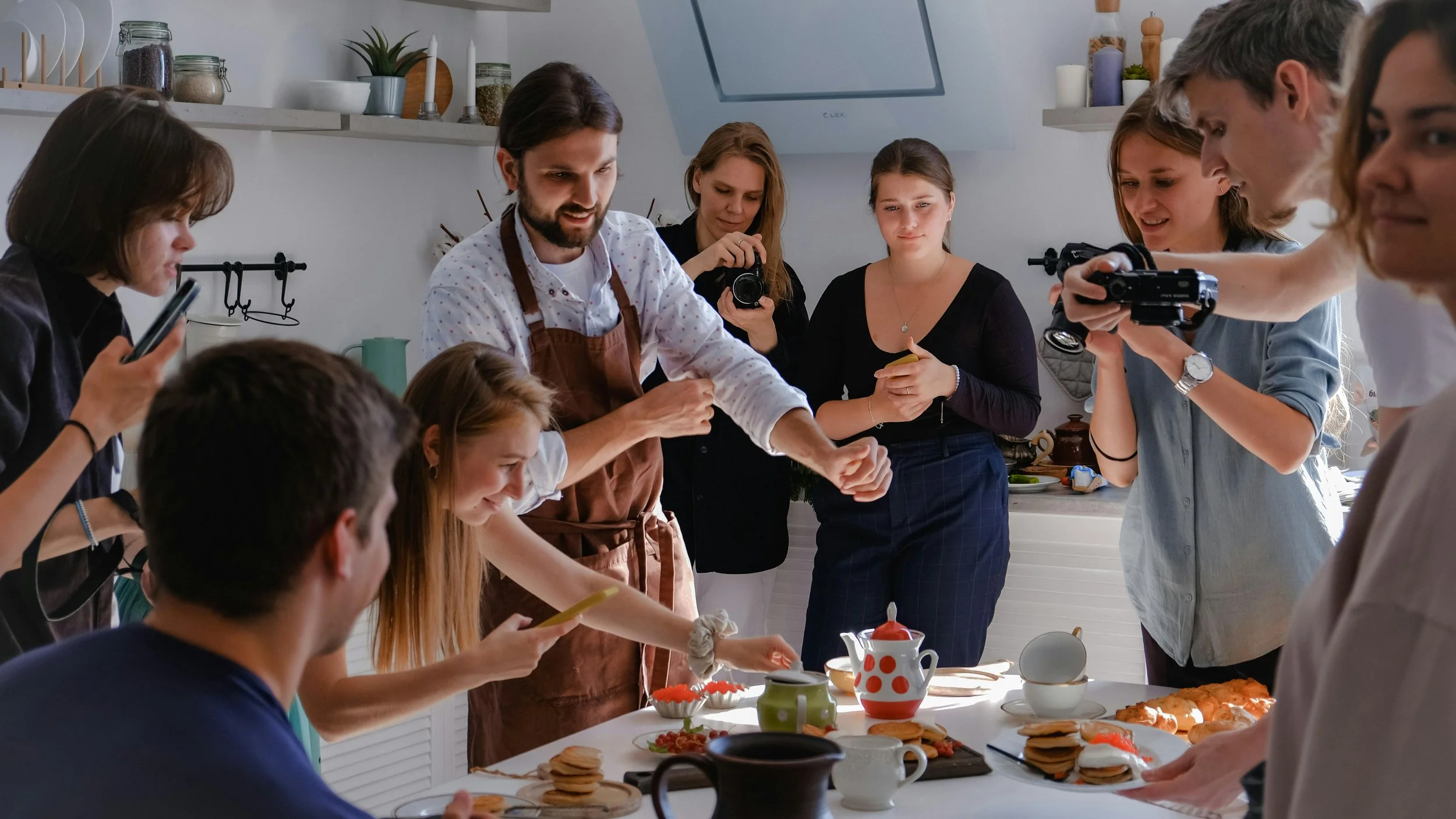 A group of people gathered around a kitchen counter, baking and decorating cookies, with plates of cookies and teapots on the table.
