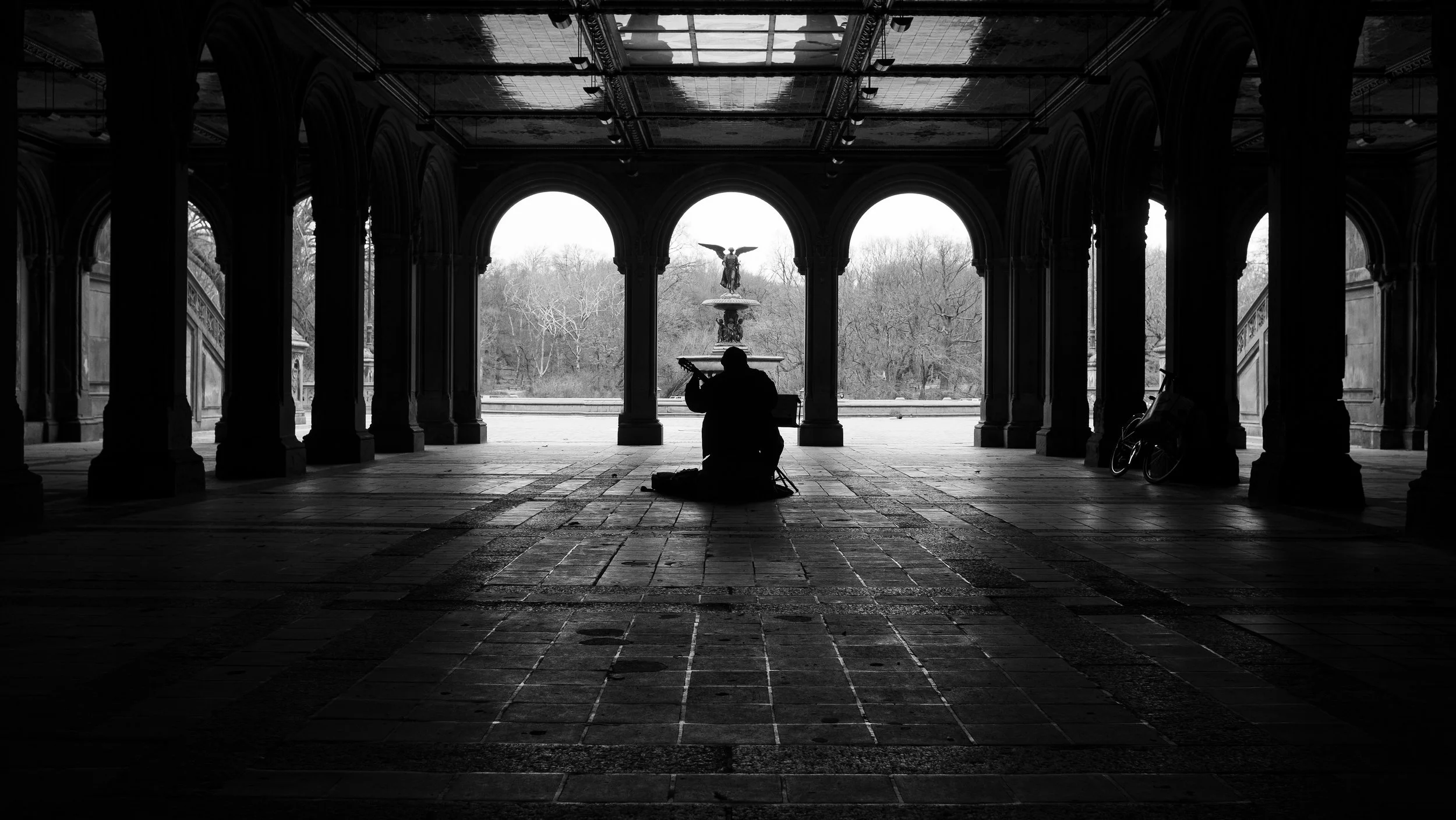 Silhouette of a person sitting on the ground, playing a guitar, in a covered outdoor structure with arches, columns, and a fountain in the background. Black and white photo.