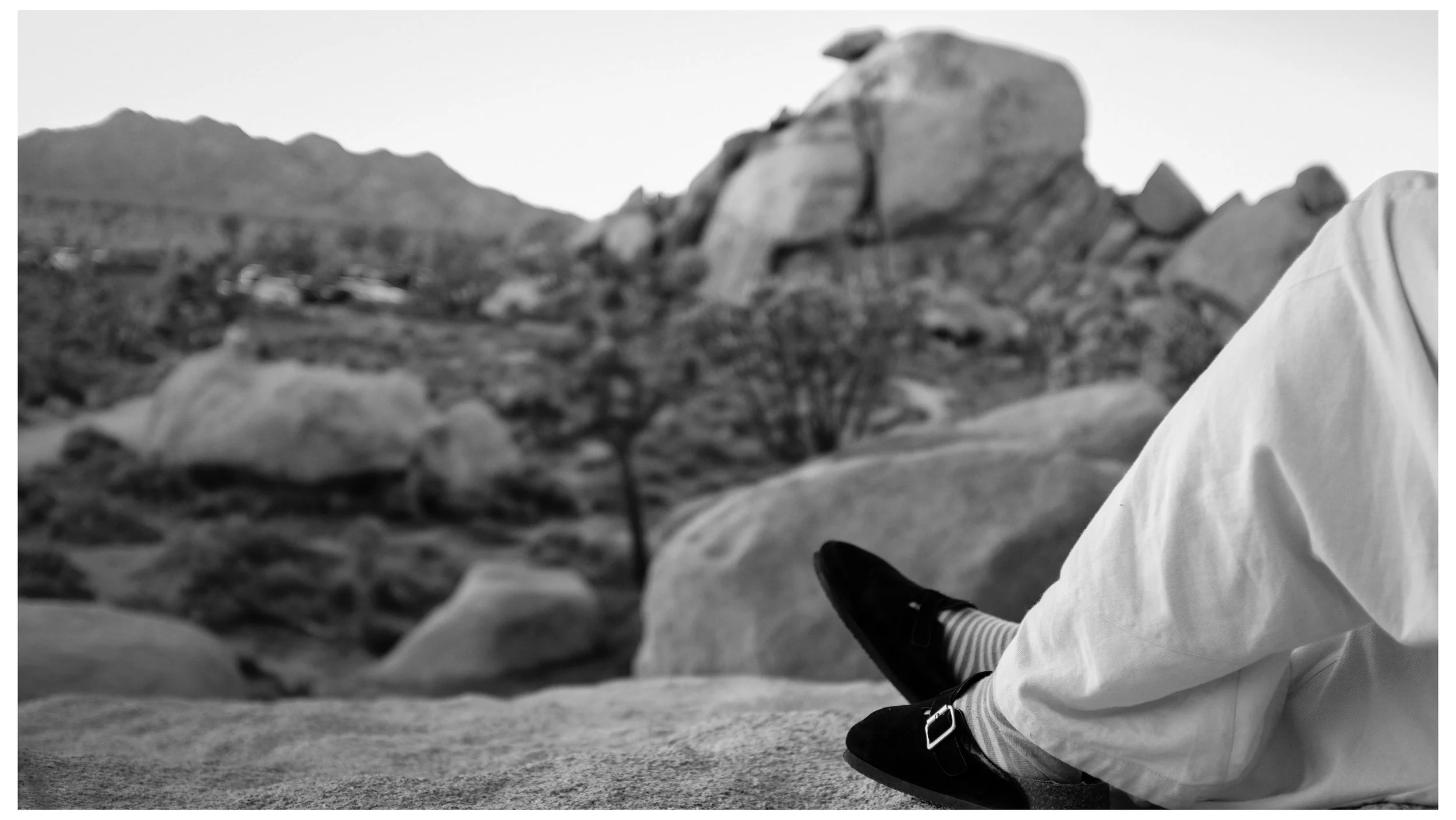 Close-up of a person's lower body and shoes, sitting on rocky terrain with mountains in the background.