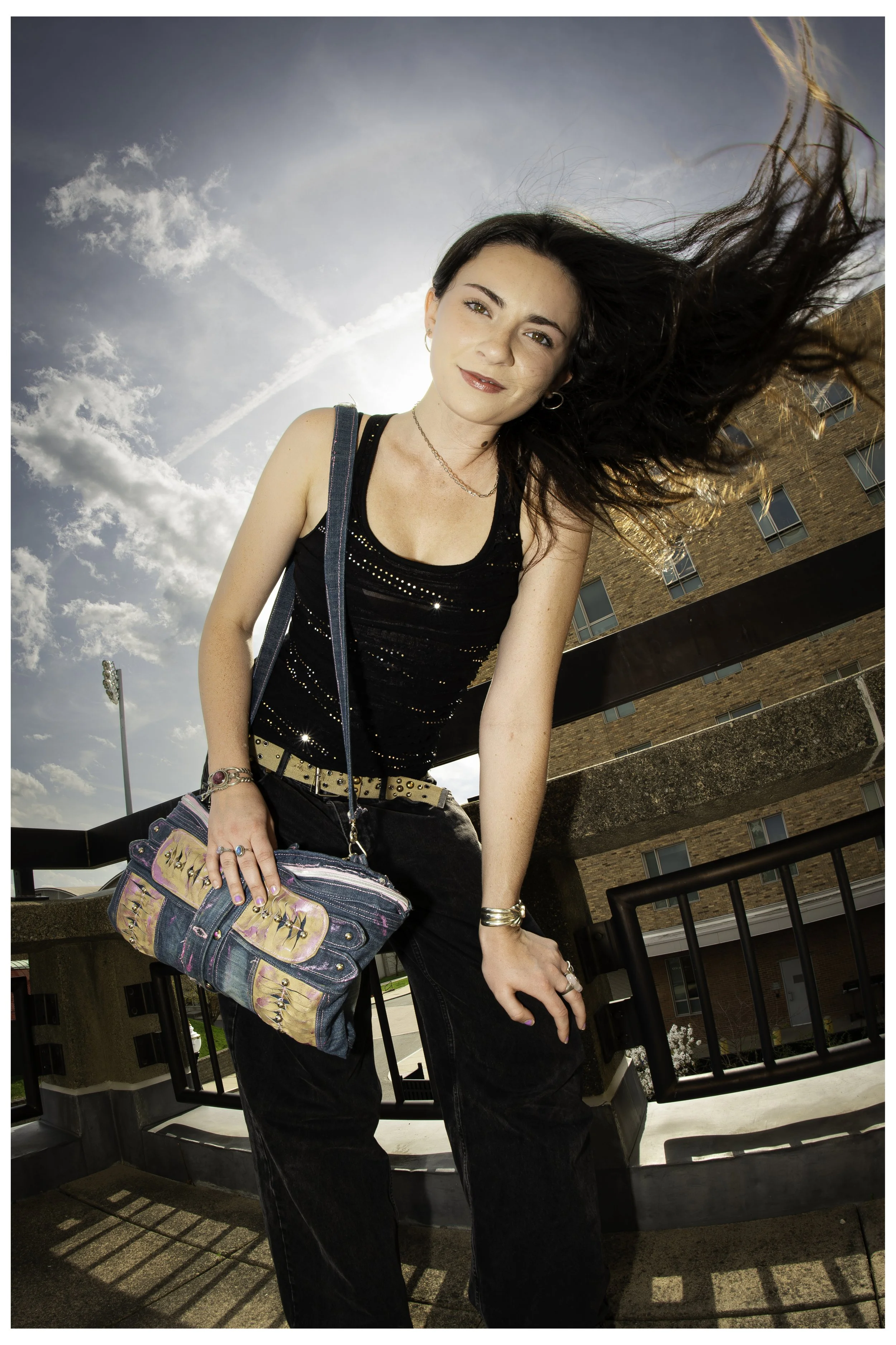 Young woman with long dark hair, smiling, standing outdoors on a sunny day, wearing a black tank top with shiny details, black pants, accessorized with jewelry, holding a large colorful bag, in front of a building with windows and a railing.