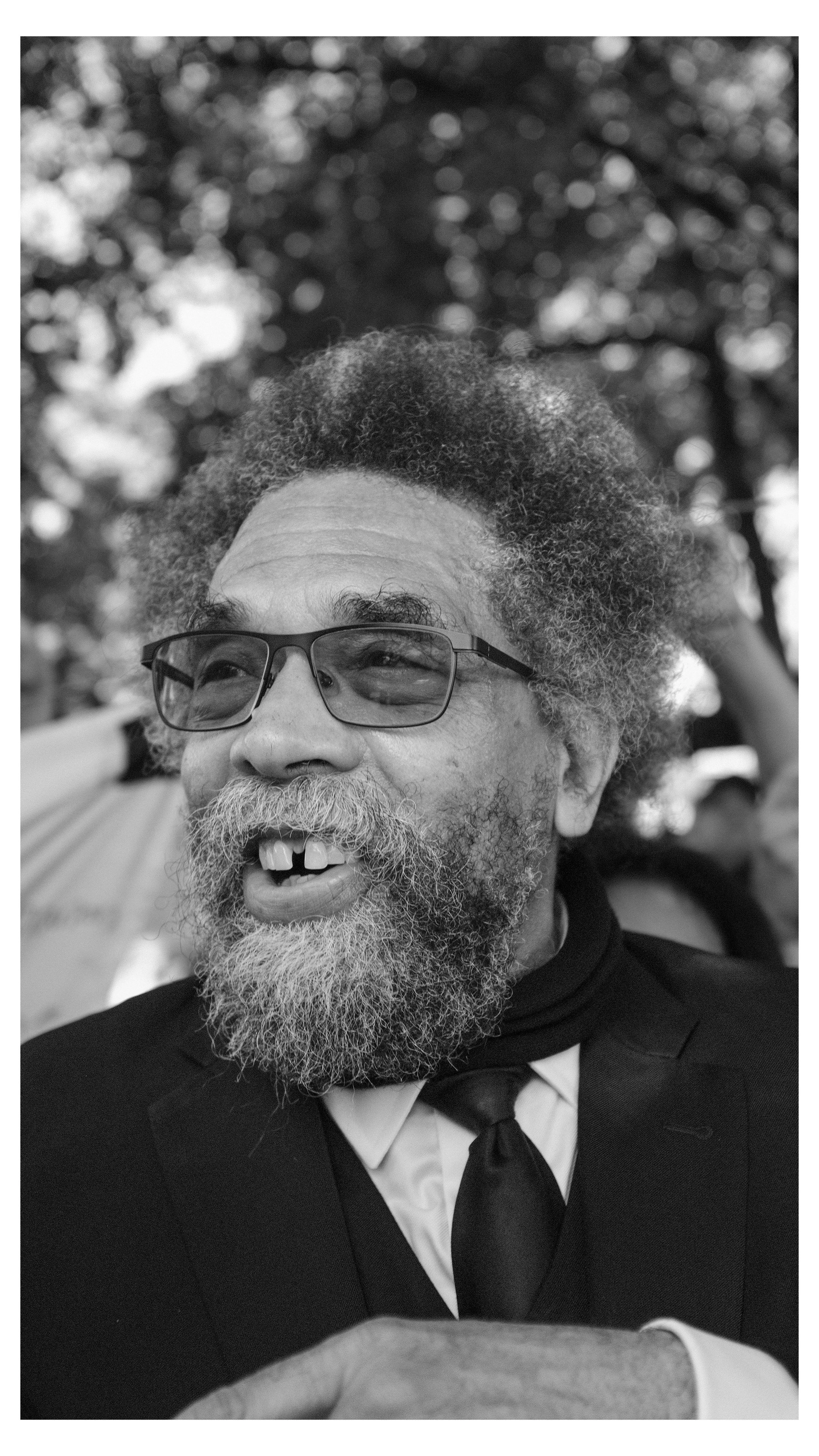 A black and white photo of a smiling man with glasses, gray beard, and missing teeth, wearing a tuxedo and bow tie. Trees are blurred in the background.