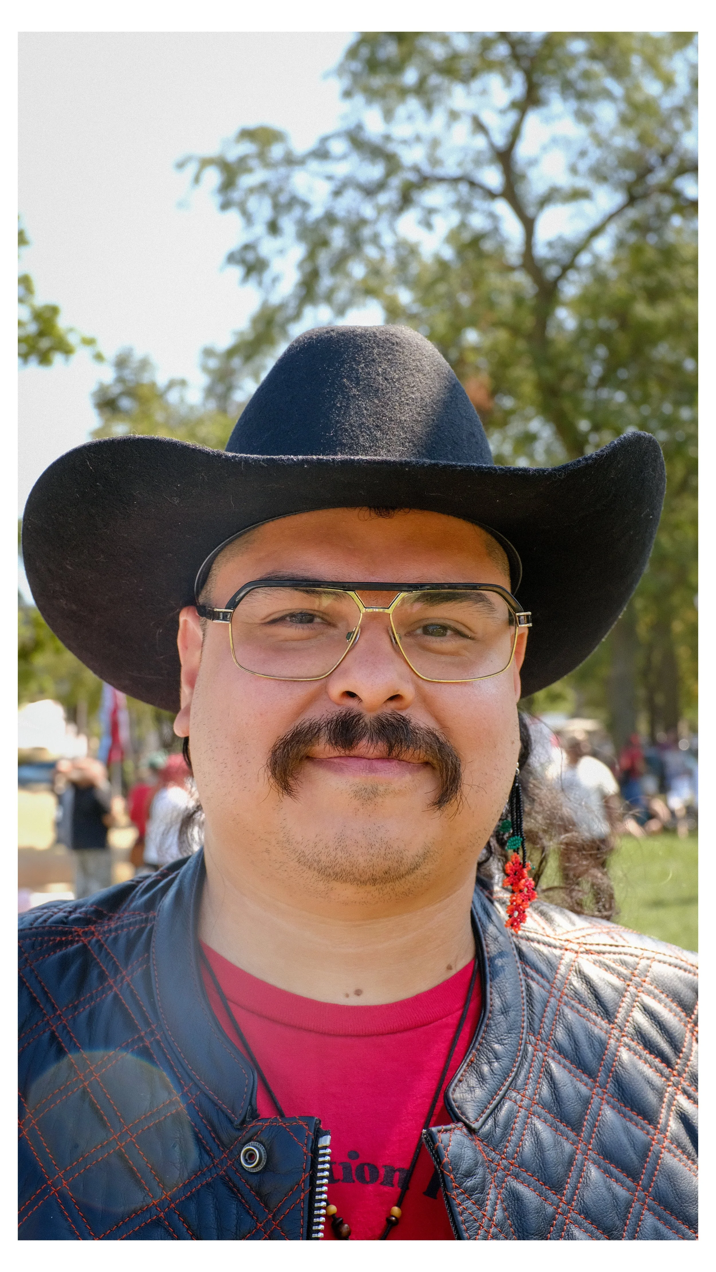 A man with glasses, a mustache, and a goatee wearing a black cowboy hat, a red shirt, and a leather jacket, smiling outdoors with trees and people in the background.