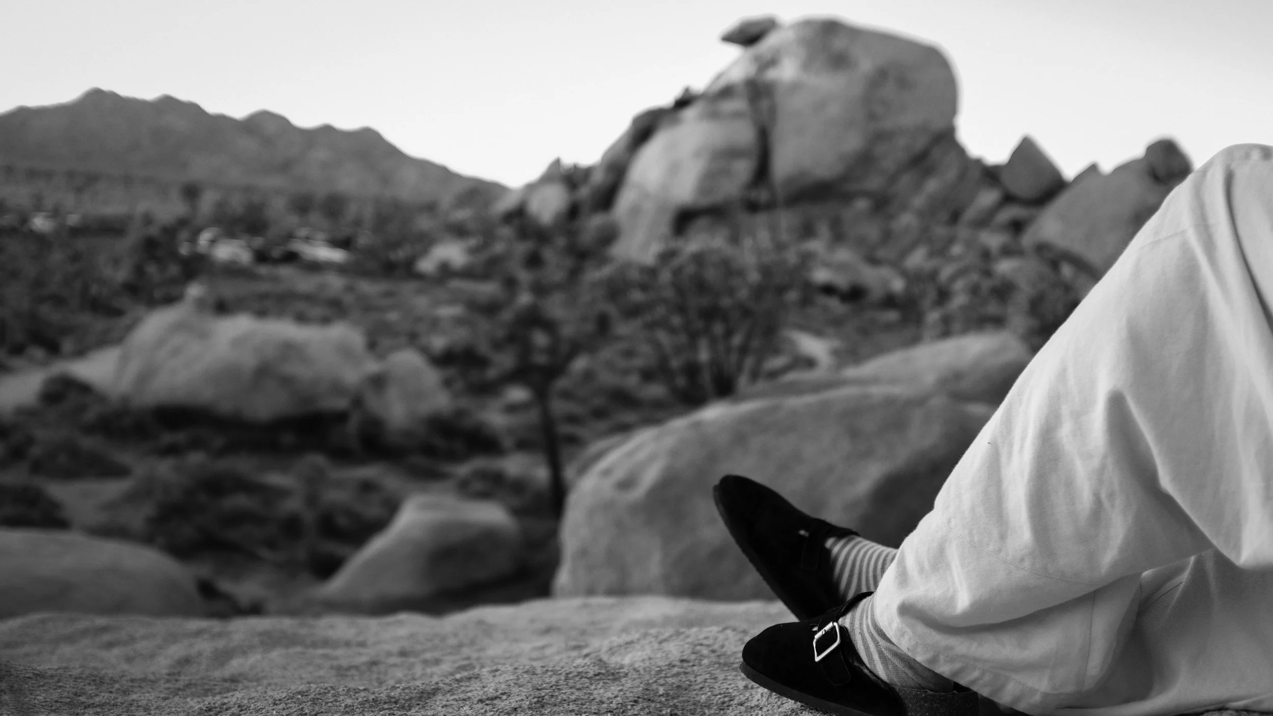 Close-up of a person sitting outdoors on rocky terrain, wearing striped socks, black shoes, and white pants, with a mountain and desert landscape in the background.