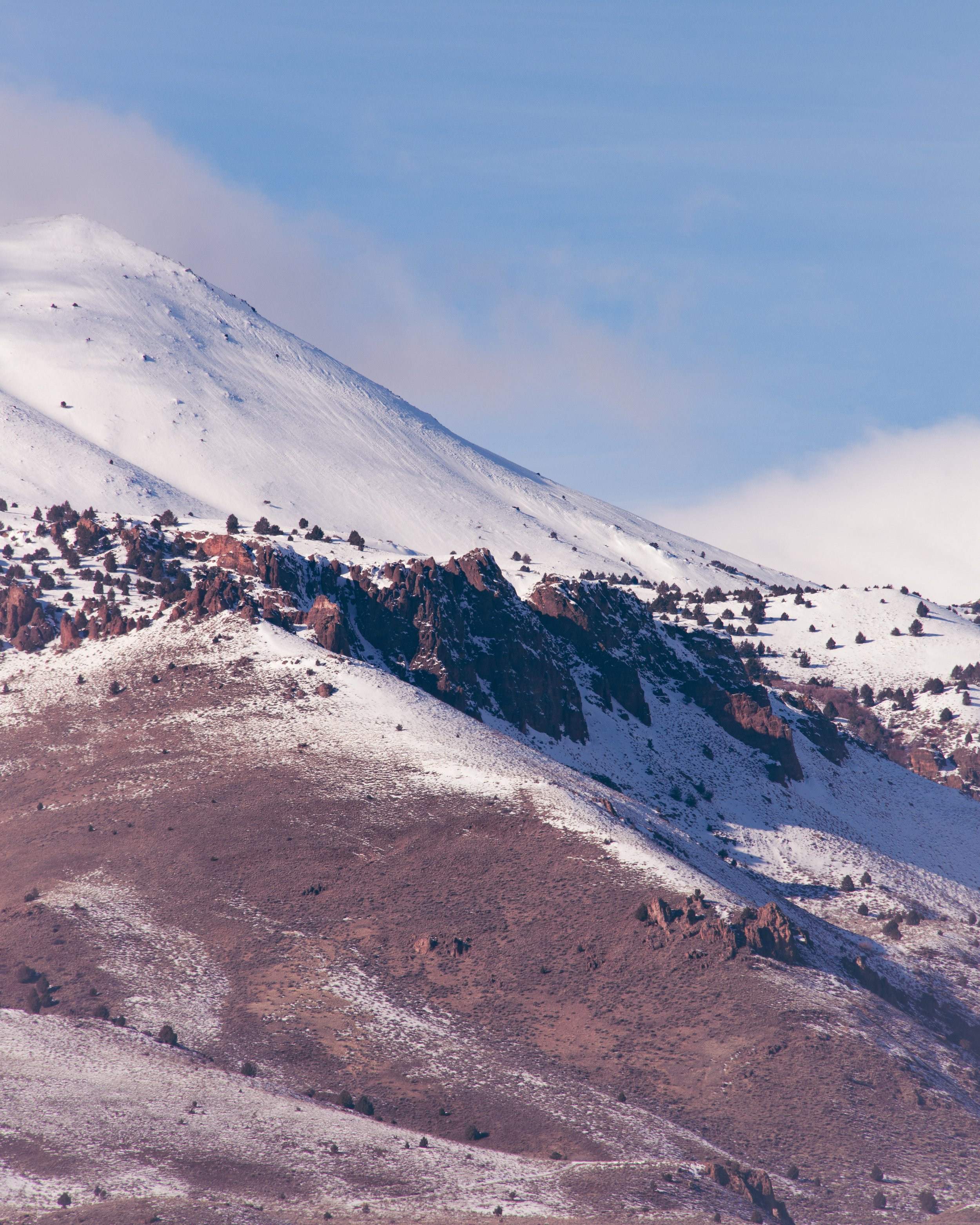 Snow-covered mountain with rocky slopes and sparse vegetation, under a partly cloudy sky.