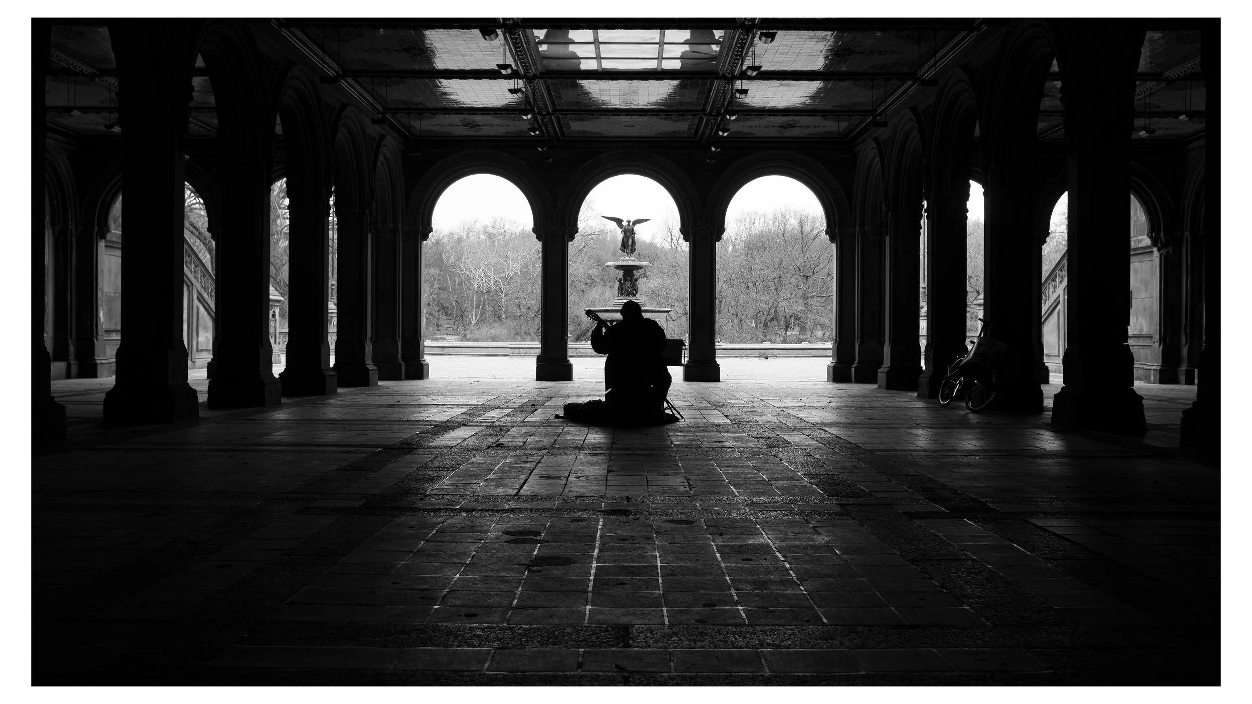 A person sitting cross-legged on the ground playing a guitar beneath a colonnade, with a fountain and trees visible in the background.