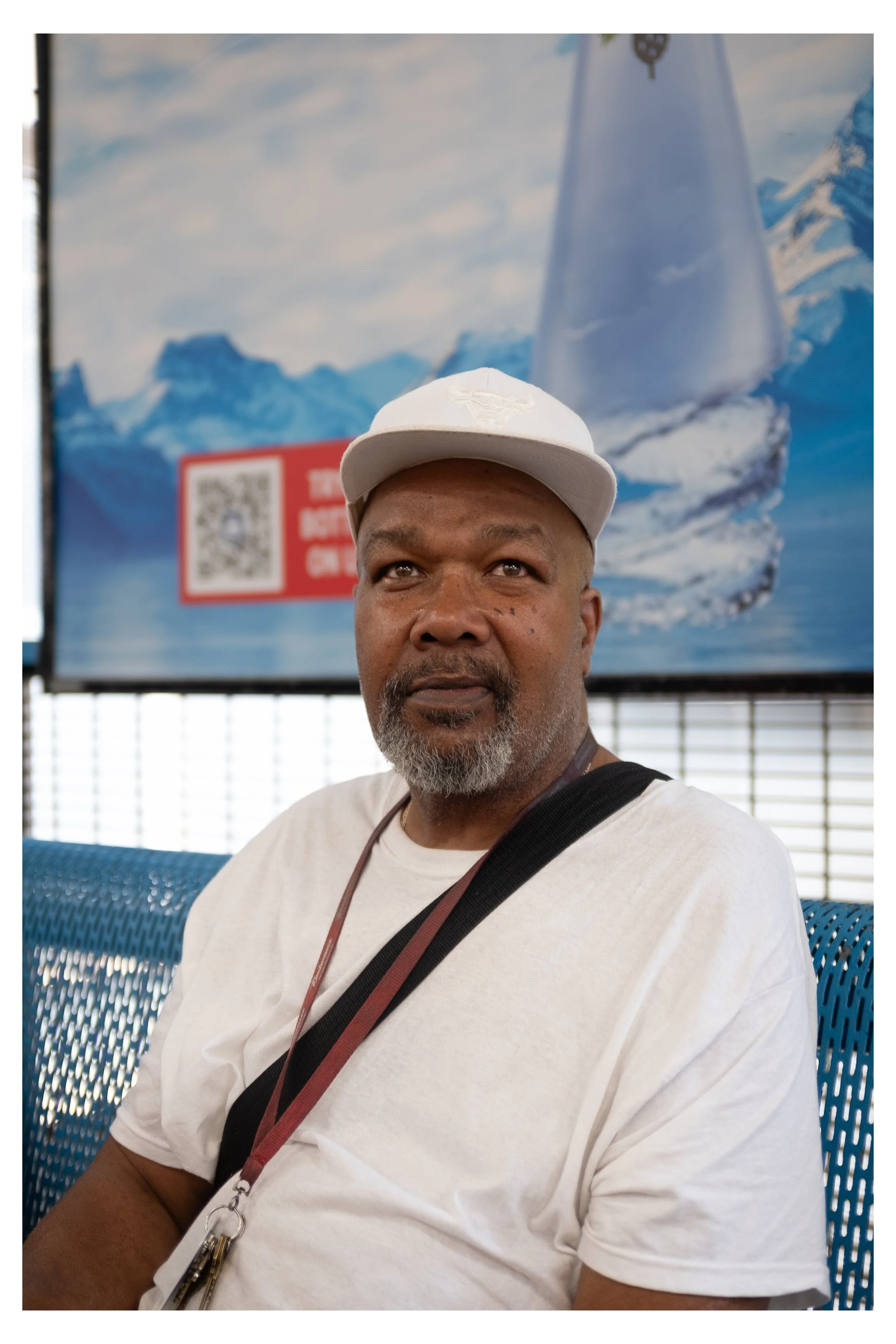 A man with a gray beard wearing a white cap and white T-shirt, sitting outdoors against a backdrop of blue sky, mountains, and a large poster with snow and ice images.