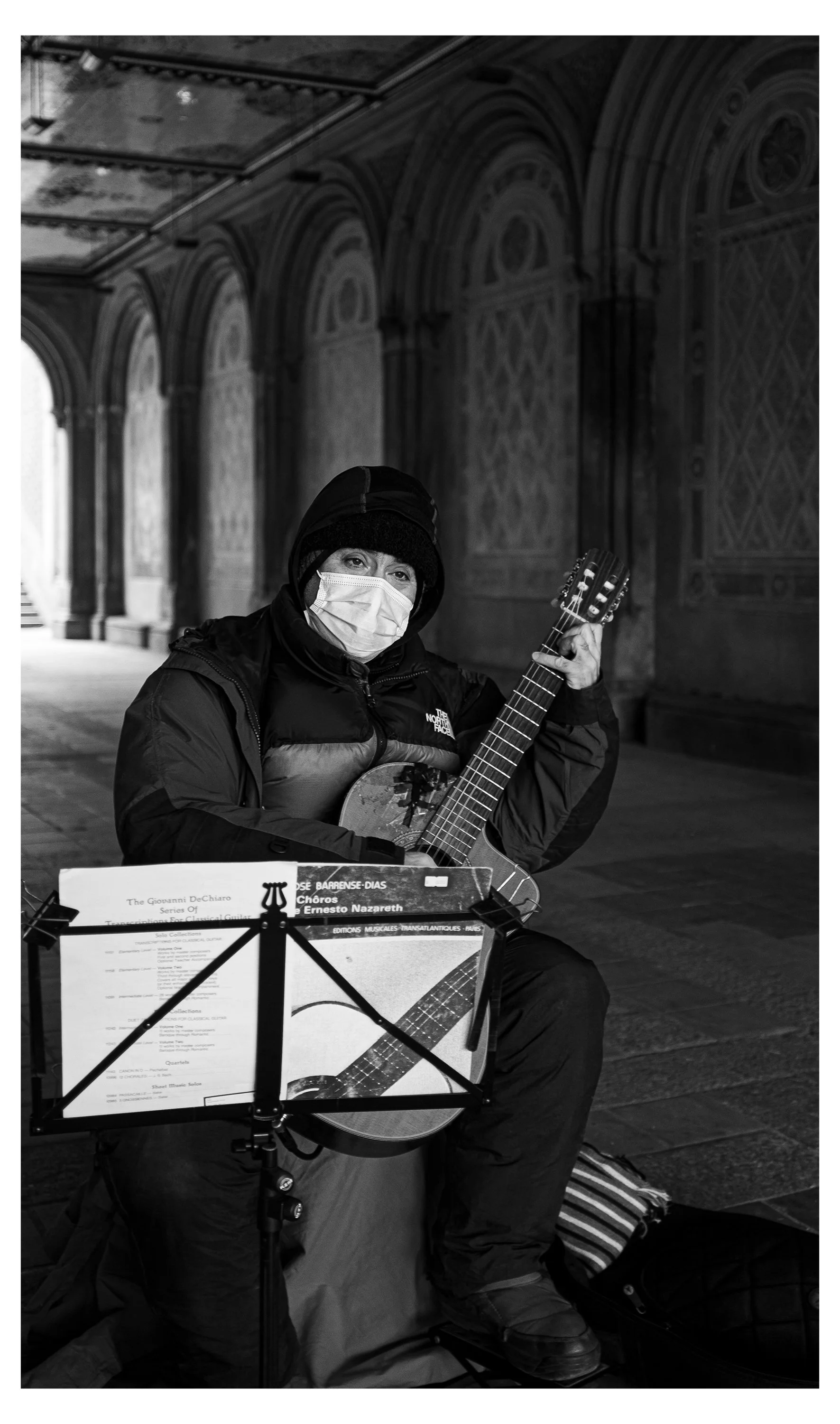 A person wearing a face mask and winter clothing playing an acoustic guitar, sitting outdoors with a music stand and sheet music in front of them, in a dimly lit architectural setting.
