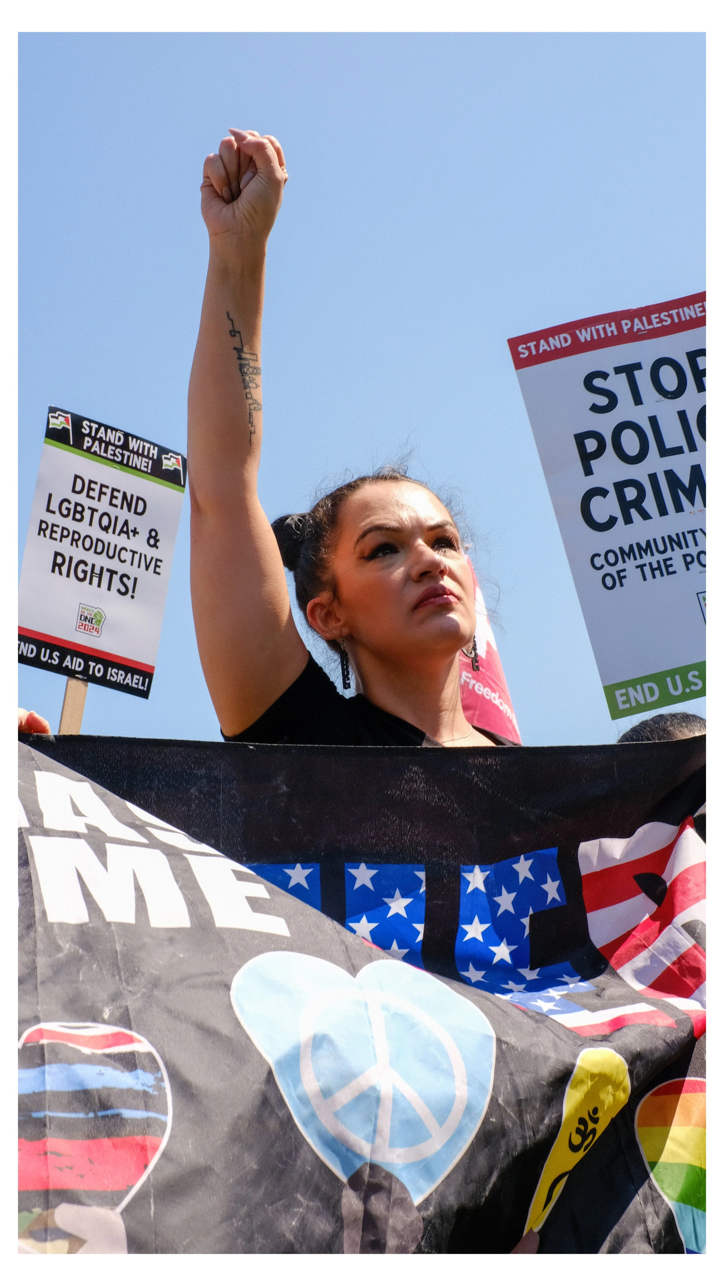 A woman with raised fist participating in a protest or rally, holding signs advocating for Palestine, LGBTQIA+ rights, and social justice, with a banner showing peace and unity symbols.