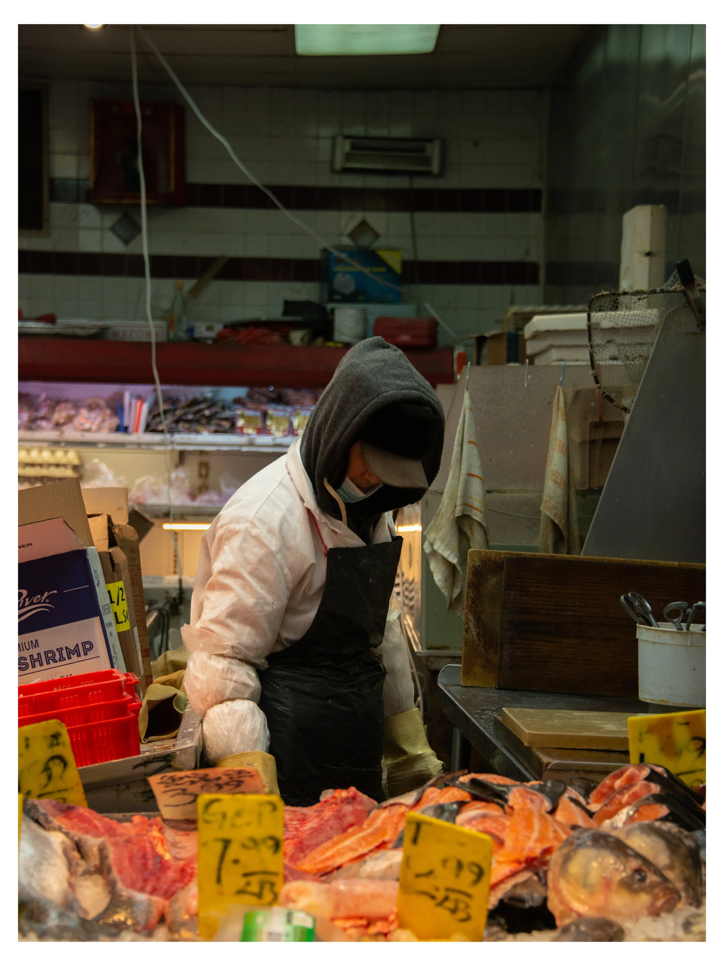 A person wearing a white coat, black apron, and gray hoodie working behind a seafood display in a market. The display includes fish and seafood on ice with yellow price tags.