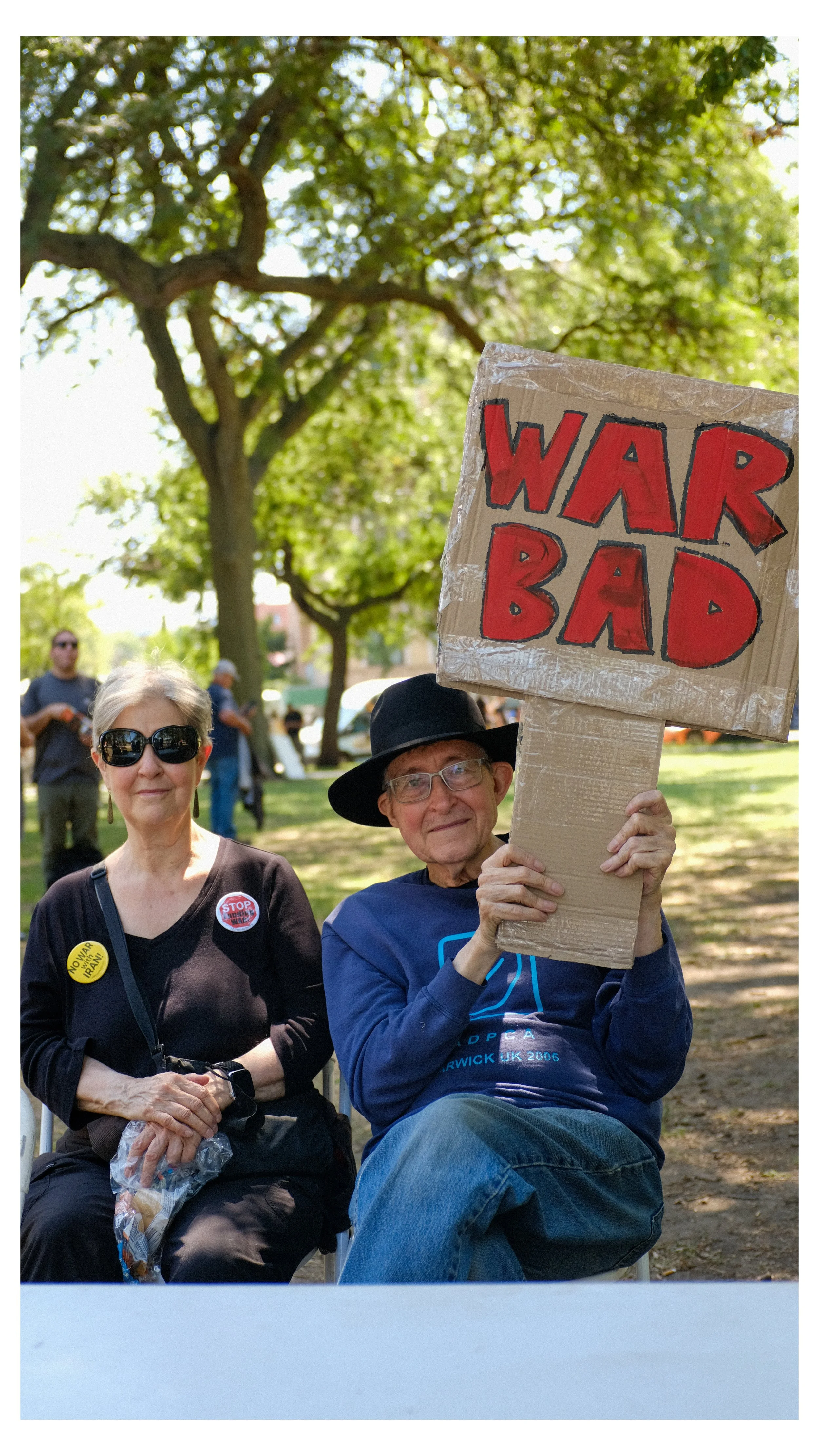 An elderly man holding a cardboard sign that reads 'WAR BAD' at a protest or demonstration in a park, with a woman sitting beside him and other people and trees visible in the background.