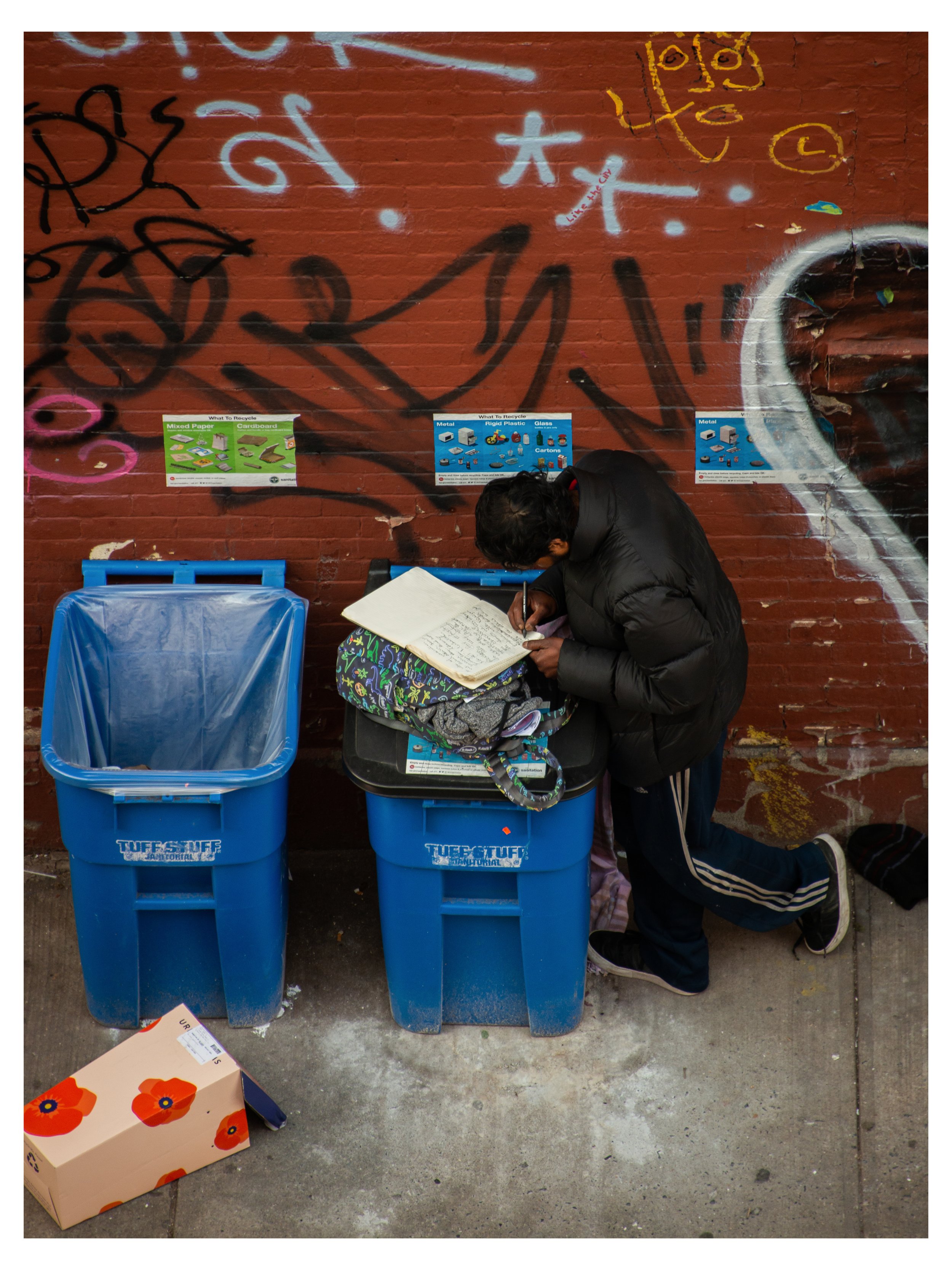 A person is kneeling next to three blue recycling bins labeled 'Turf's Turf' with a brick wall painted with graffiti in the background. The person is writing or drawing in a notebook on their lap. There are posters on the wall that show different cat