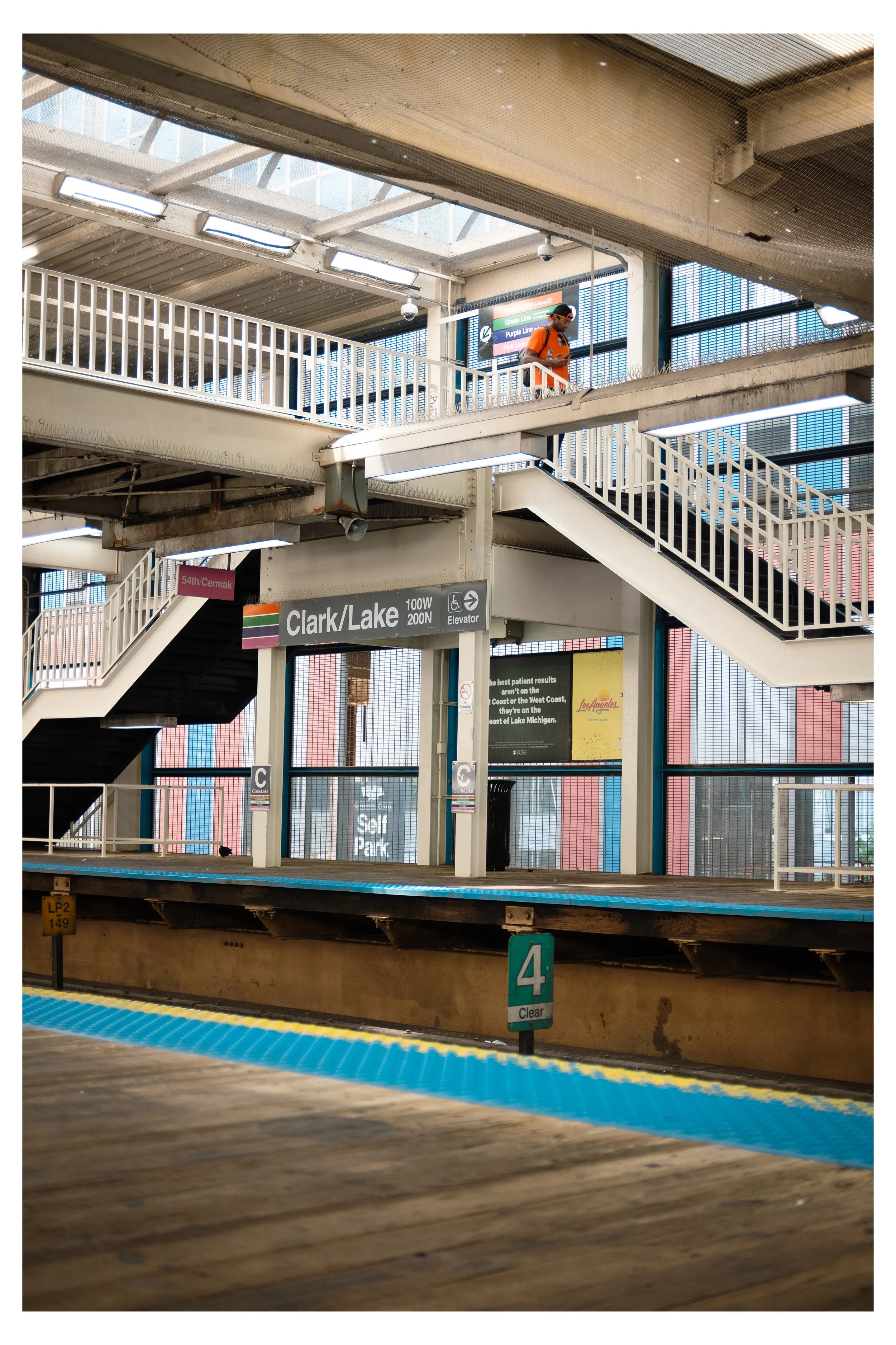 Inside a train station, a person wearing an orange shirt and black cap is walking on an upper level. The station features multiple levels, stairs with white railings, and signs indicating directions to Clark/Lake, and an elevator. Sunlight filters th