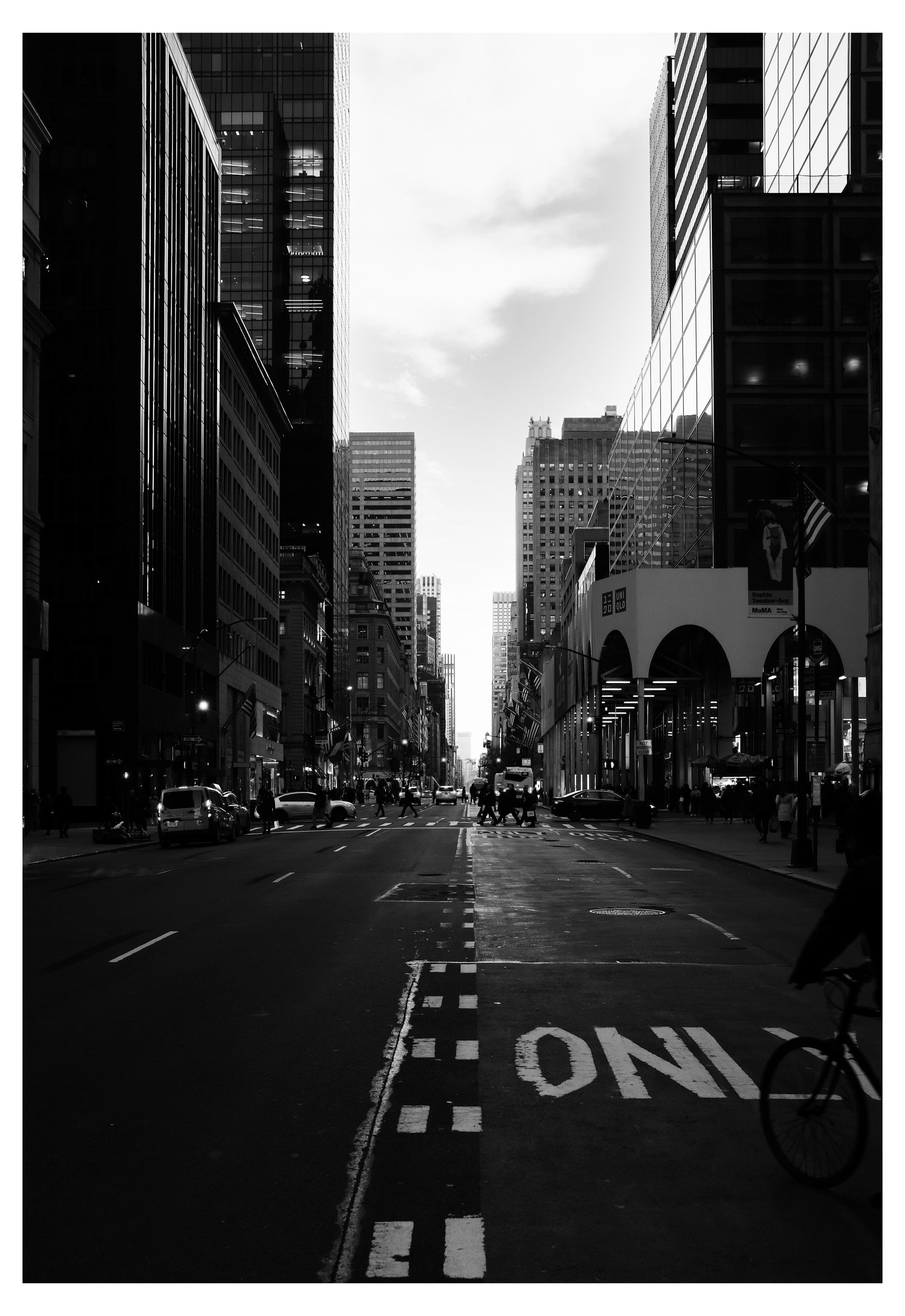 Black and white photo of a city street with tall buildings on both sides, some with reflective glass windows. There are pedestrians crossing at the intersection, cars parked along the street, and a bicycle partially visible in the lower right corner.