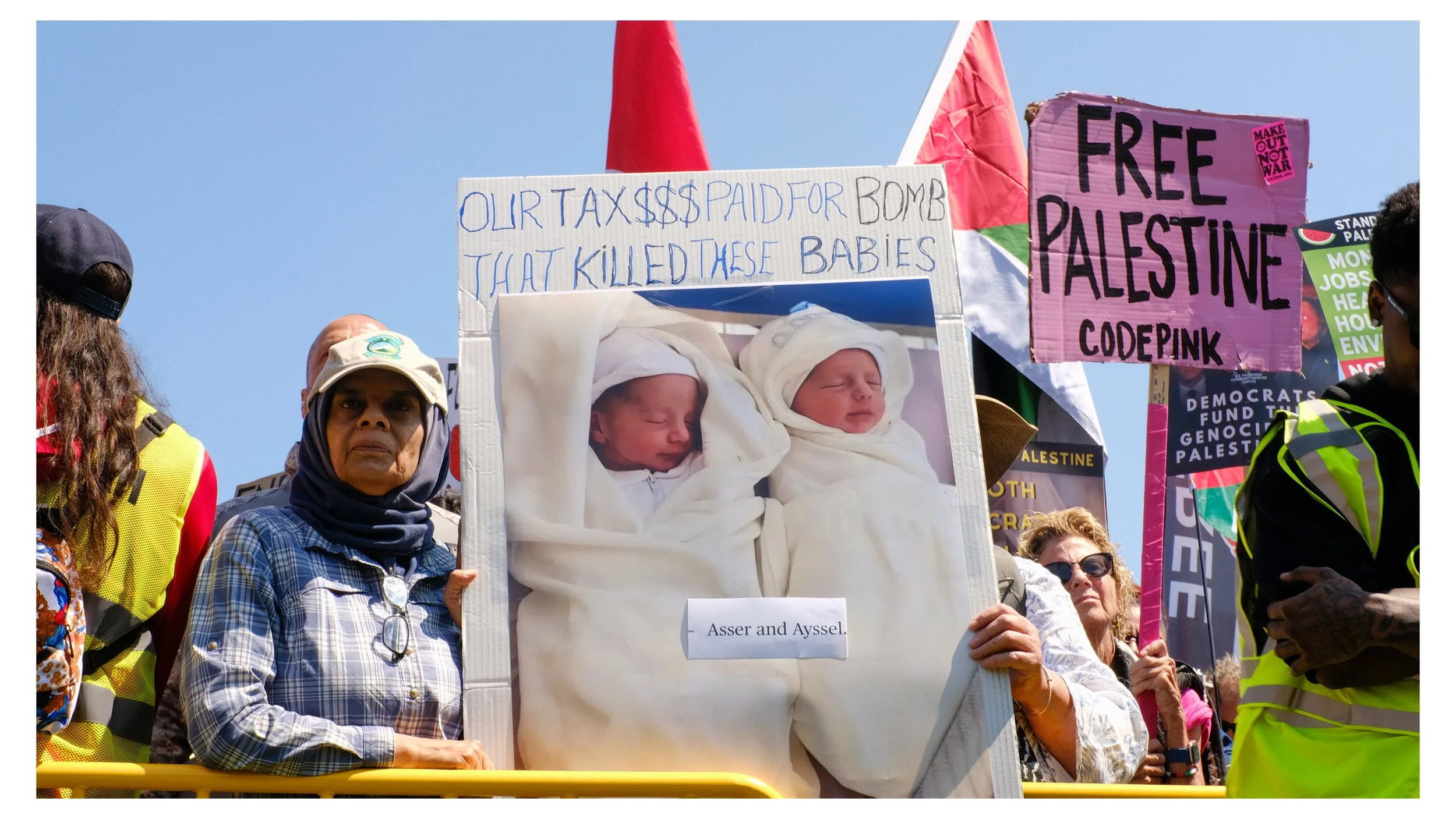 Protesters holding signs including one with a photo of newborn babies wrapped in blankets and a message about taxes paid for bombs and innocent victims, with other signs advocating for Palestinian rights and social justice.
