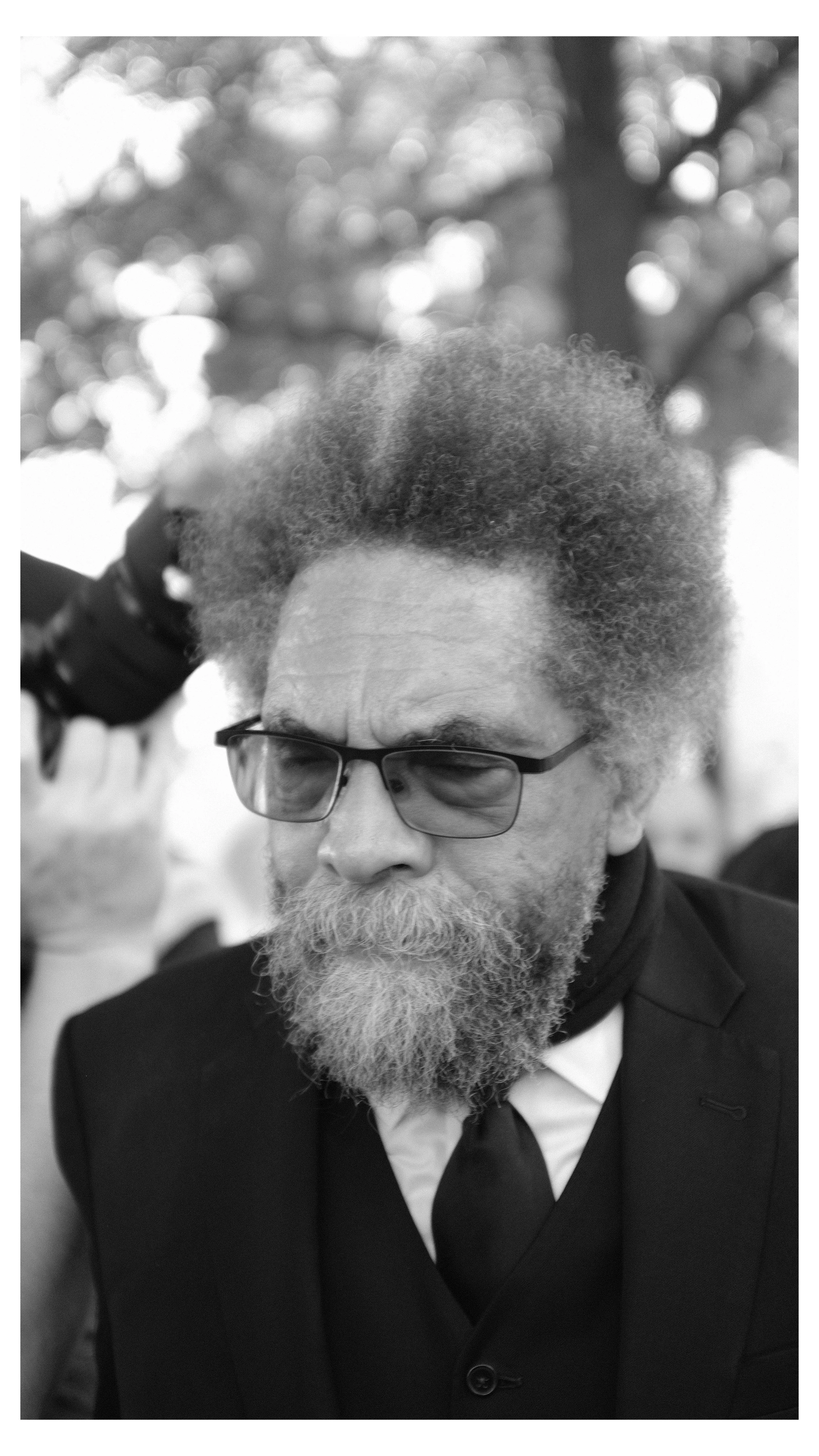 A black-and-white photo of a man with curly, gray hair, wearing glasses, a beard, and a mustache, dressed in a formal suit and tie, with a blurred background of trees and bokeh light.