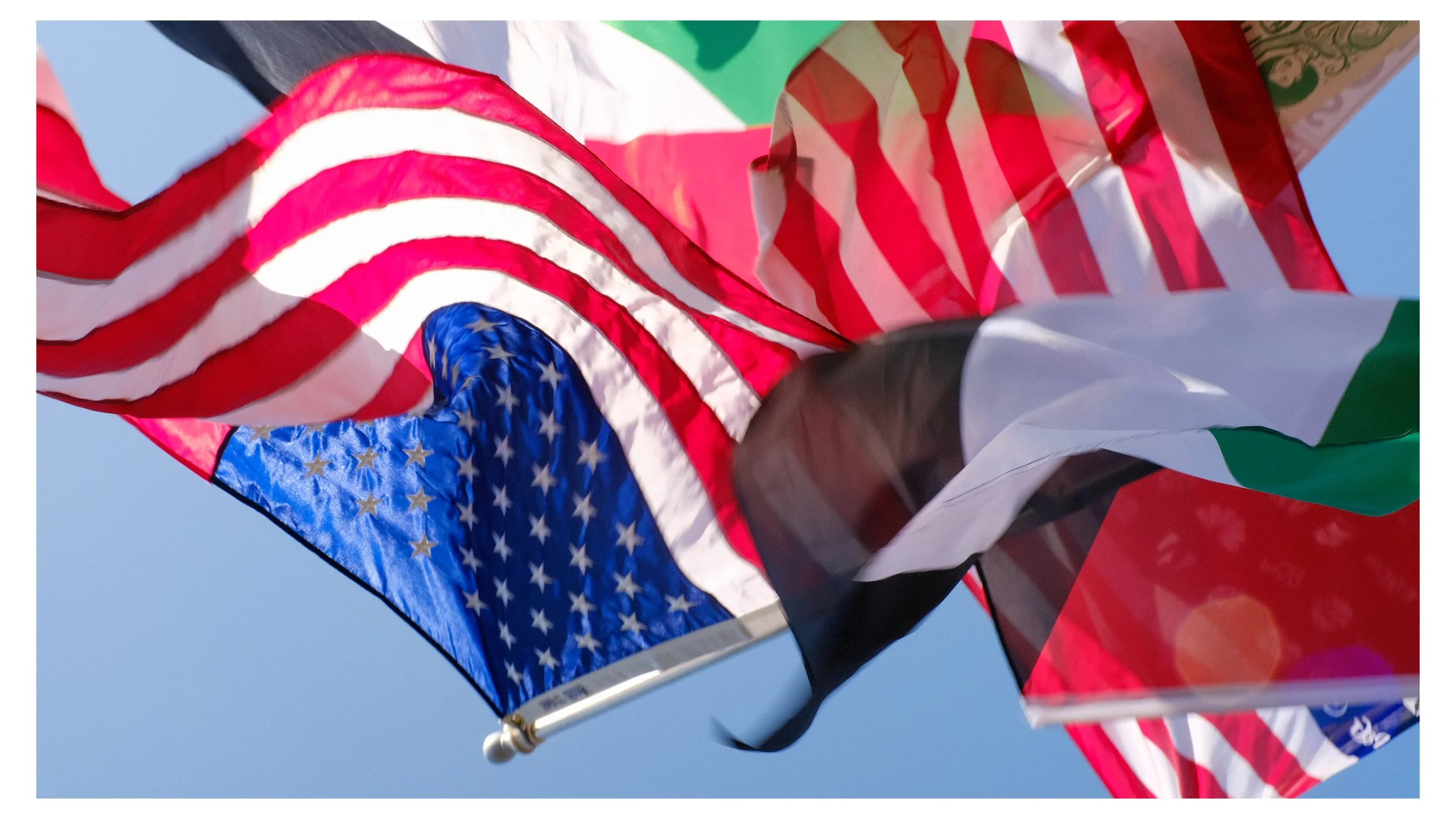 Multiple flags waving in the wind, including the American flag with red and white stripes and white stars on a blue field, and other flags with red, white, green, black, and other colors against a clear blue sky.
