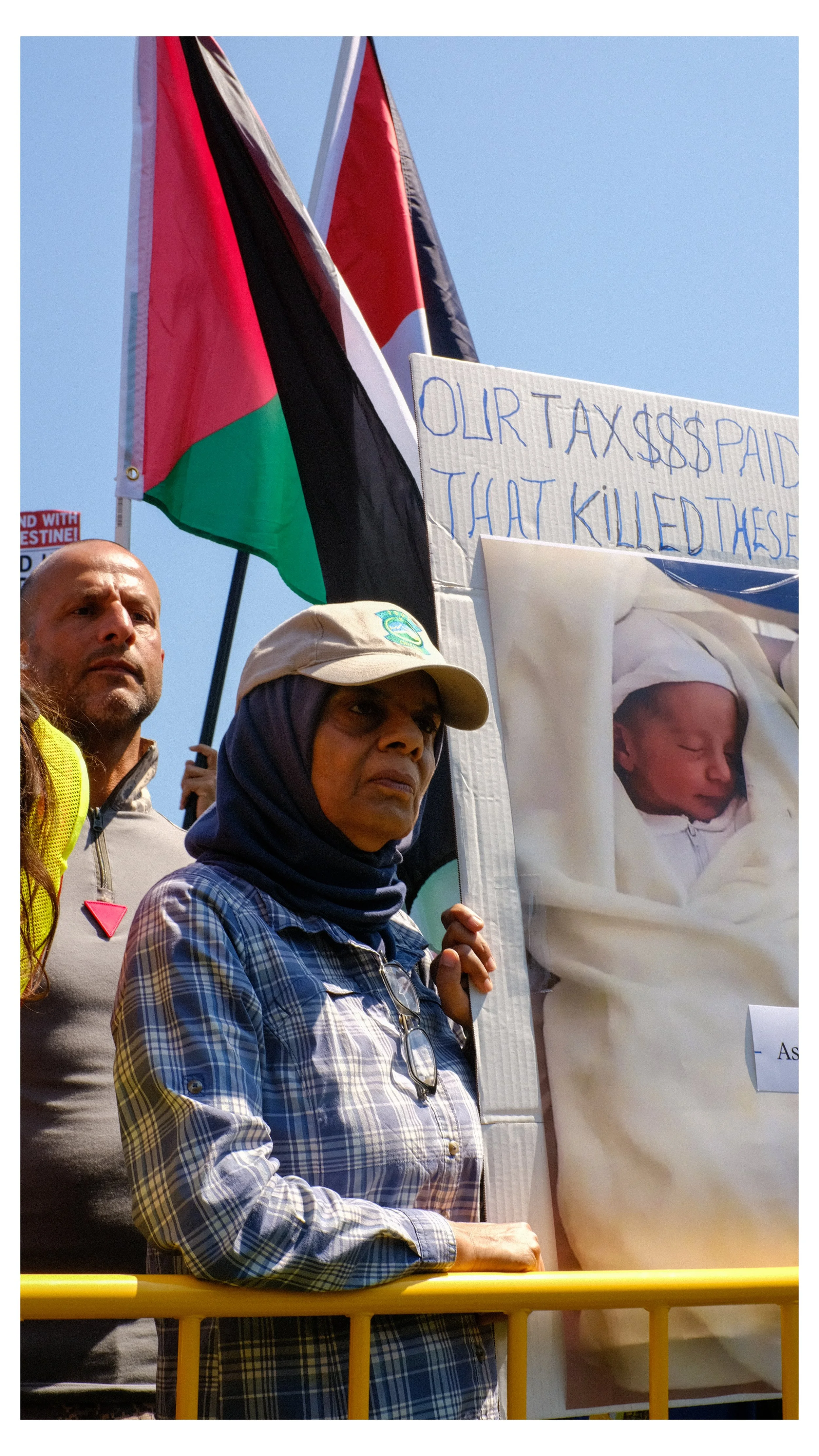 Protesters protesting against taxes, holding signs with a baby picture and a flag, standing behind a yellow barricade.
