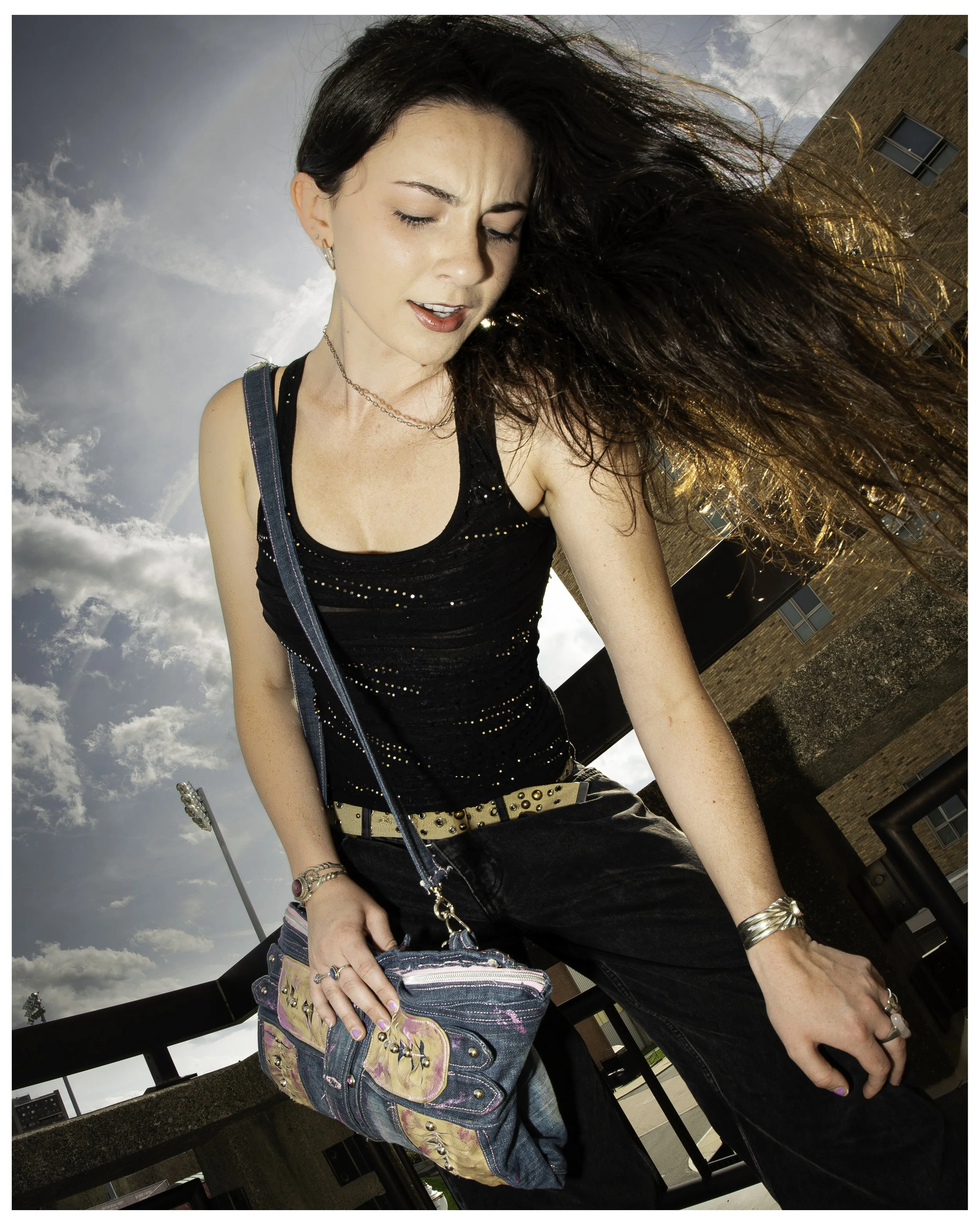 A woman with dark hair and fair skin standing outdoors on a cloudy day, wearing a black sequined tank top, black pants, various bracelets, and a beige belt with studs, holding a colorful purse, with her eyes closed and her hair blowing in the wind, i