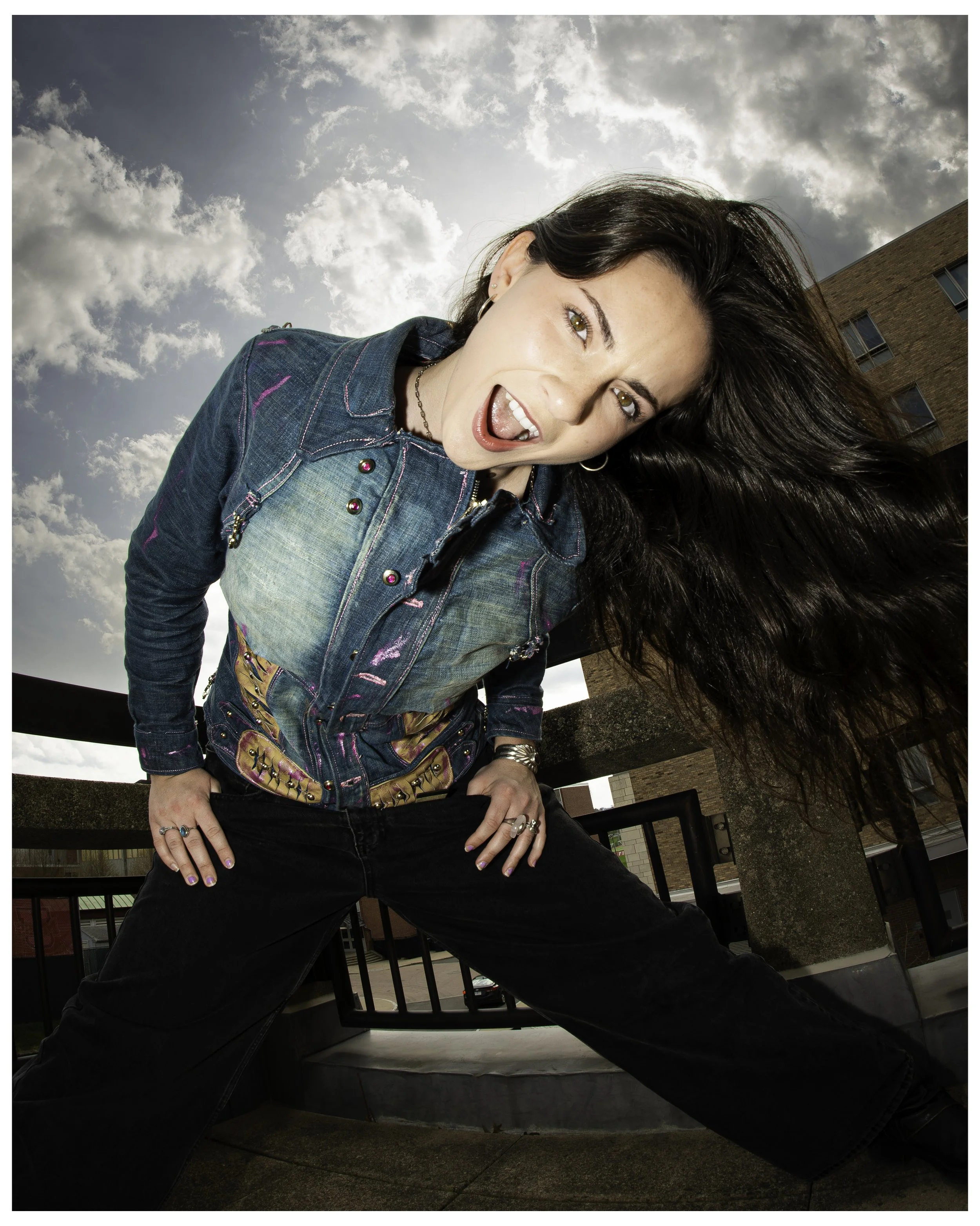 A woman with long dark hair, wearing a denim jacket and jewelry, posing with a playful expression against a cloudy sky and urban buildings in the background.