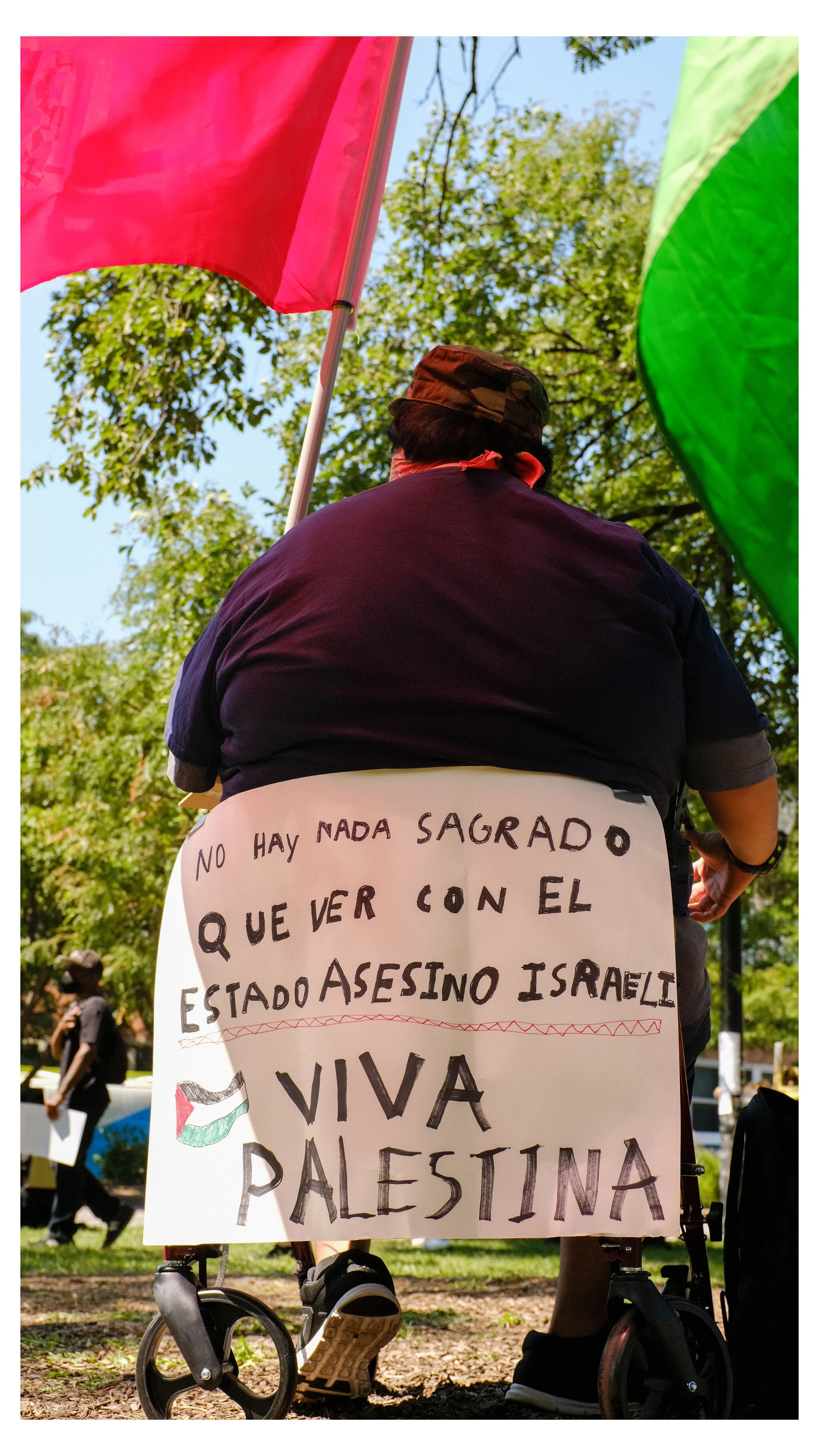 Person seated in a mobility scooter holding a protest sign in Spanish, with flags in the background, during a sunny outdoor event with trees and other protesters in the distance.