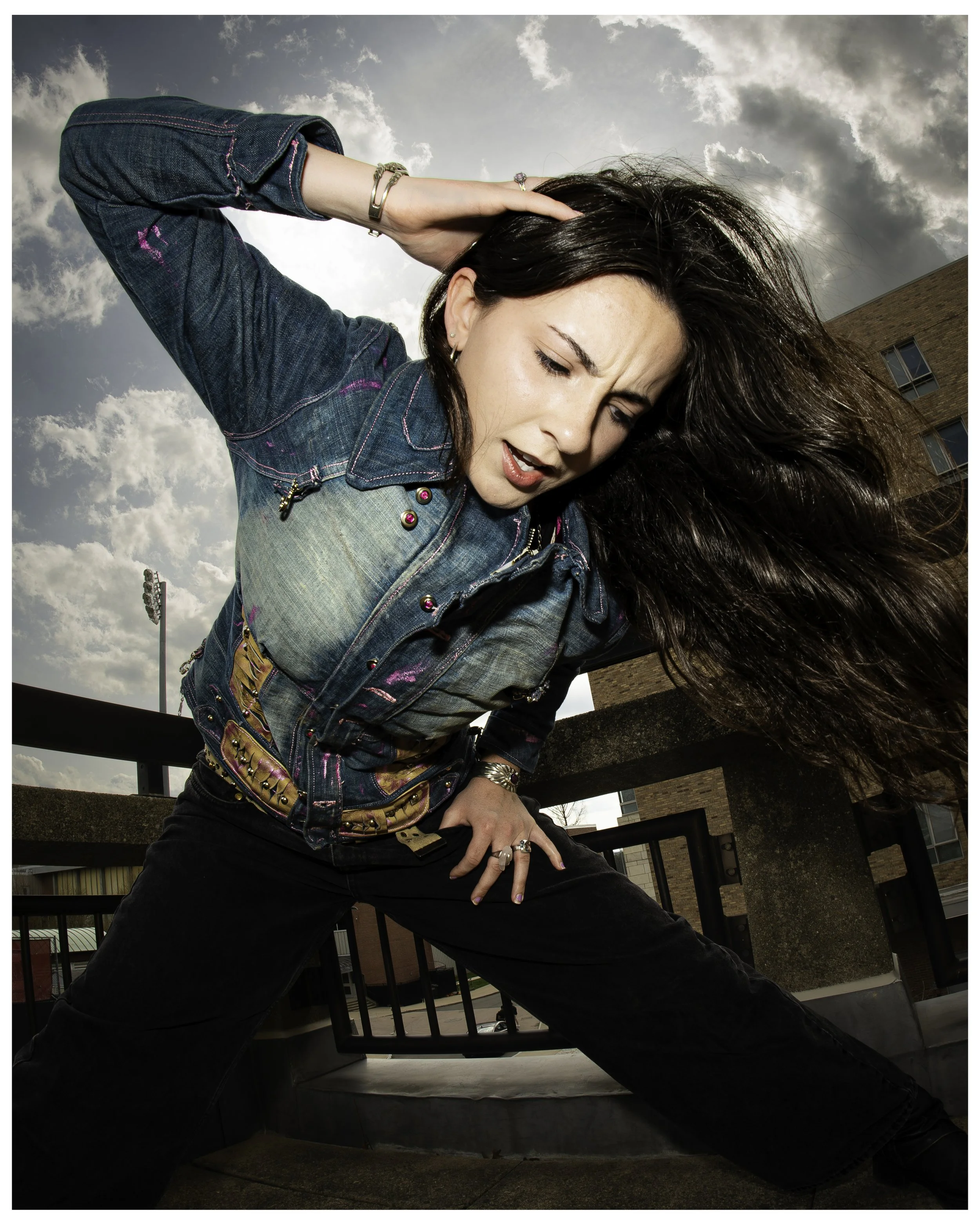 A woman in a denim jacket with pink accents and black pants is outside on a balcony, striking a dramatic pose with her head bent and one hand behind her head. Her long, dark hair is flowing and she appears to be expressing emotion or surprise. The ba