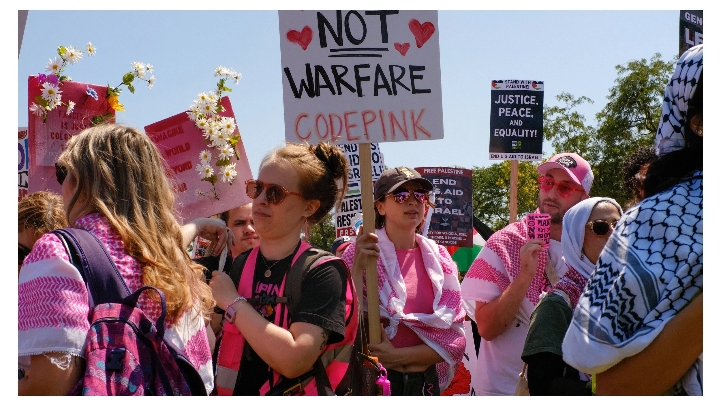 People participating in a protest march, holding signs advocating against war and supporting Palestine, with some dressed in pink and white, and others wearing headscarves and sunglasses.
