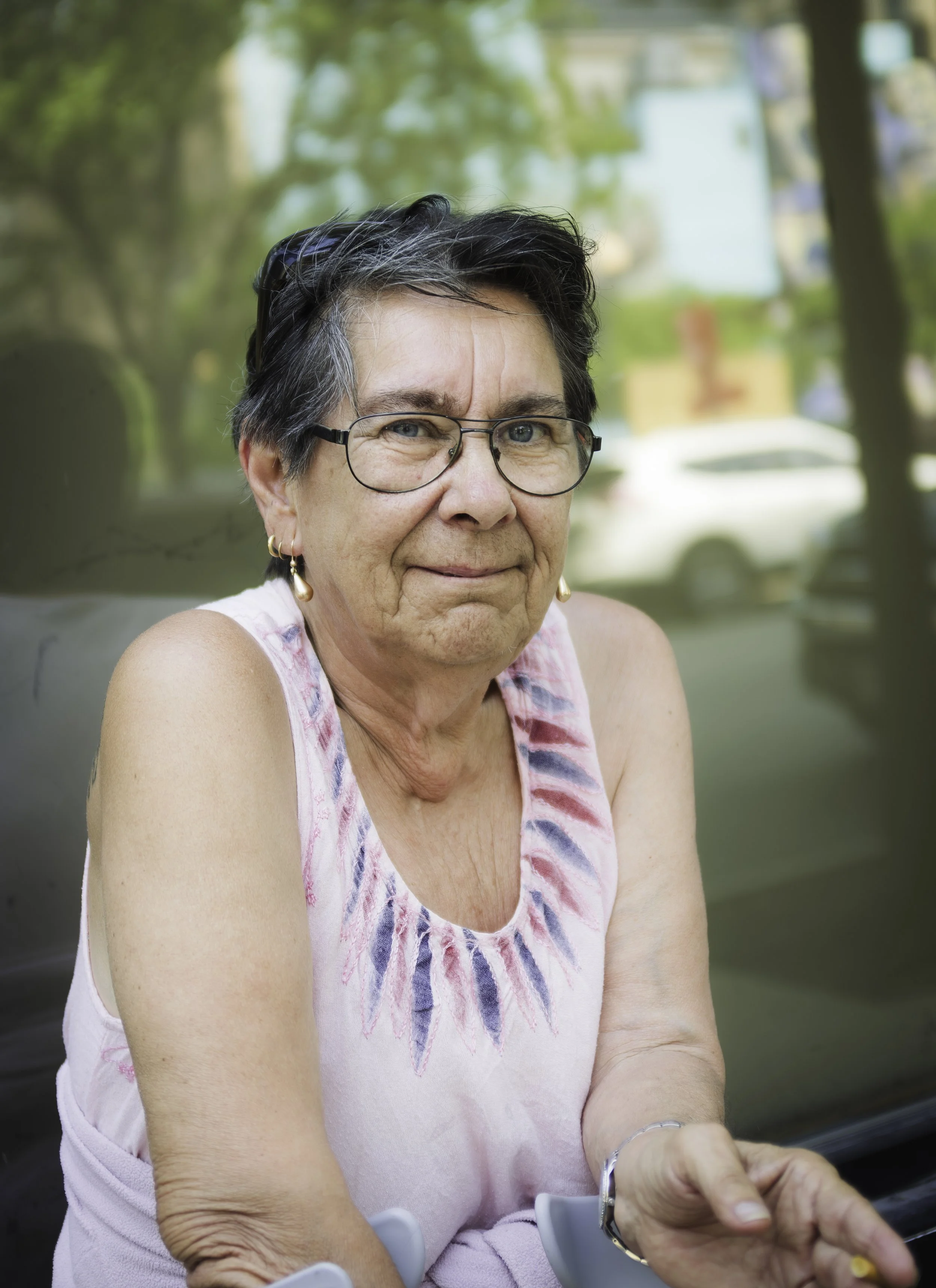 Close-up of an elderly woman with short, dark gray hair and glasses, wearing a sleeveless pink and white patterned top, sitting outdoors with a blurred green and yellow background.