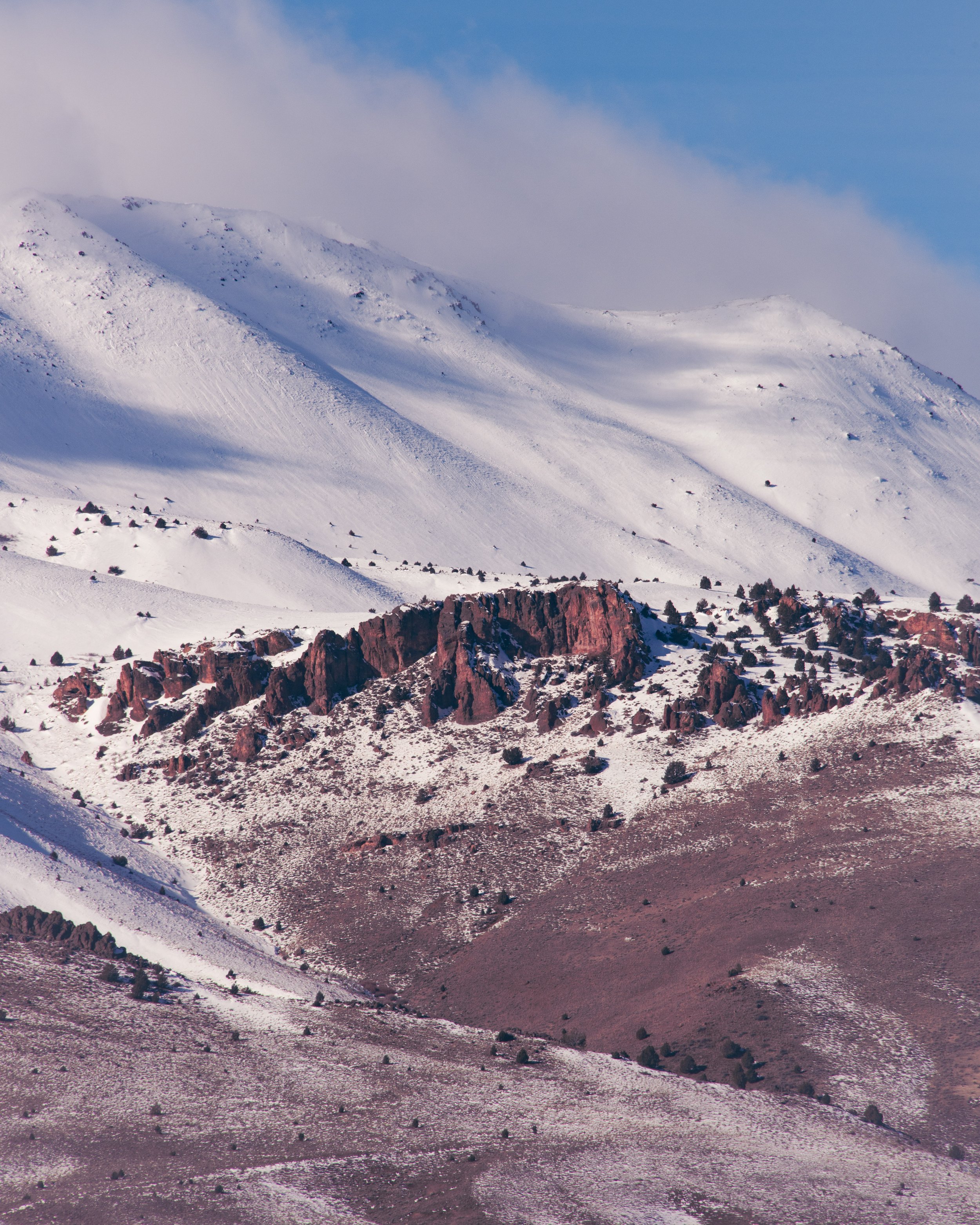 Snow-covered mountain landscape with a rocky reddish-brown cliff in the foreground and a blue sky with some clouds in the background.