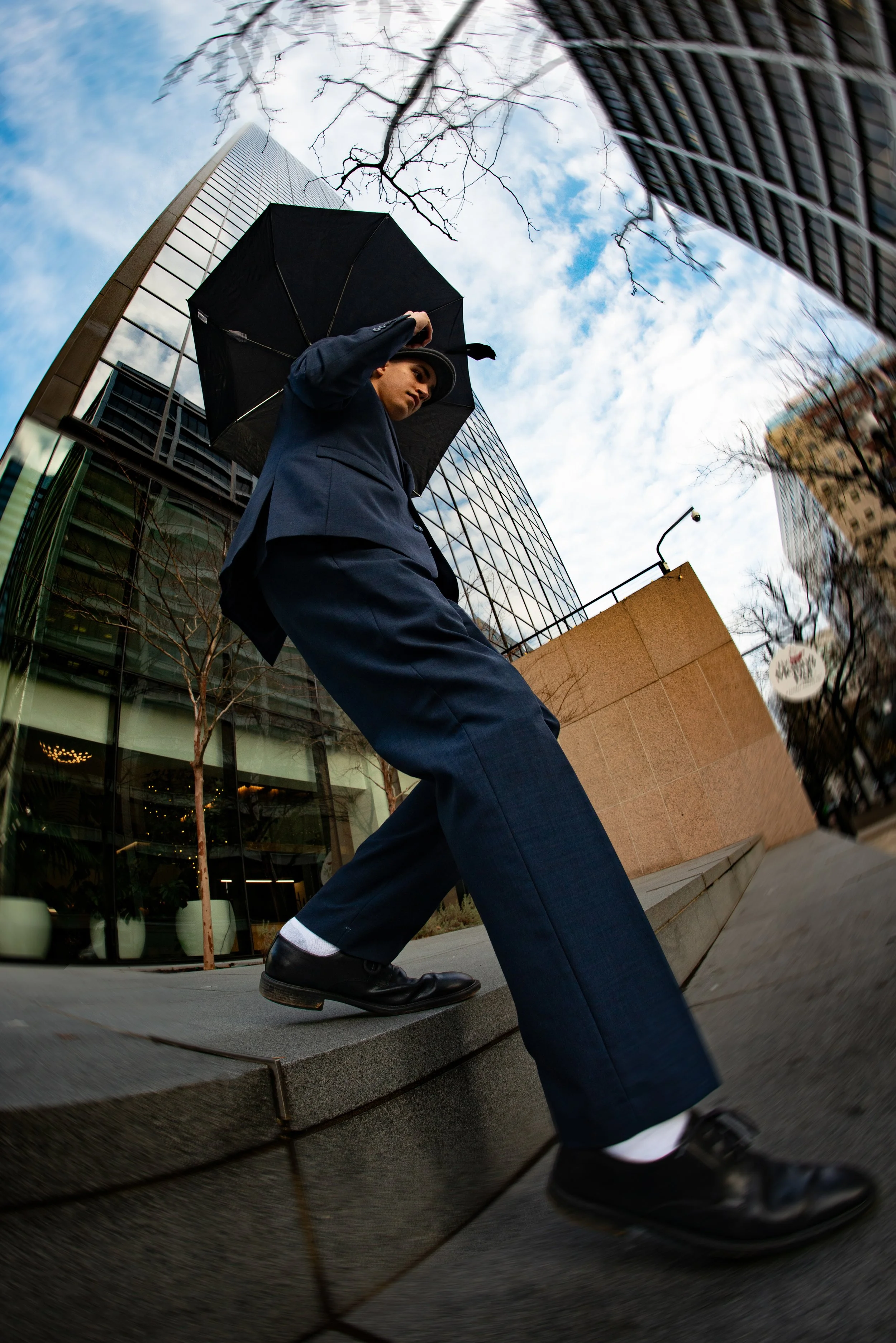 A person walking outdoors holding a black umbrella, reflected in a curved glass window with modern buildings and a partly cloudy sky in the background.