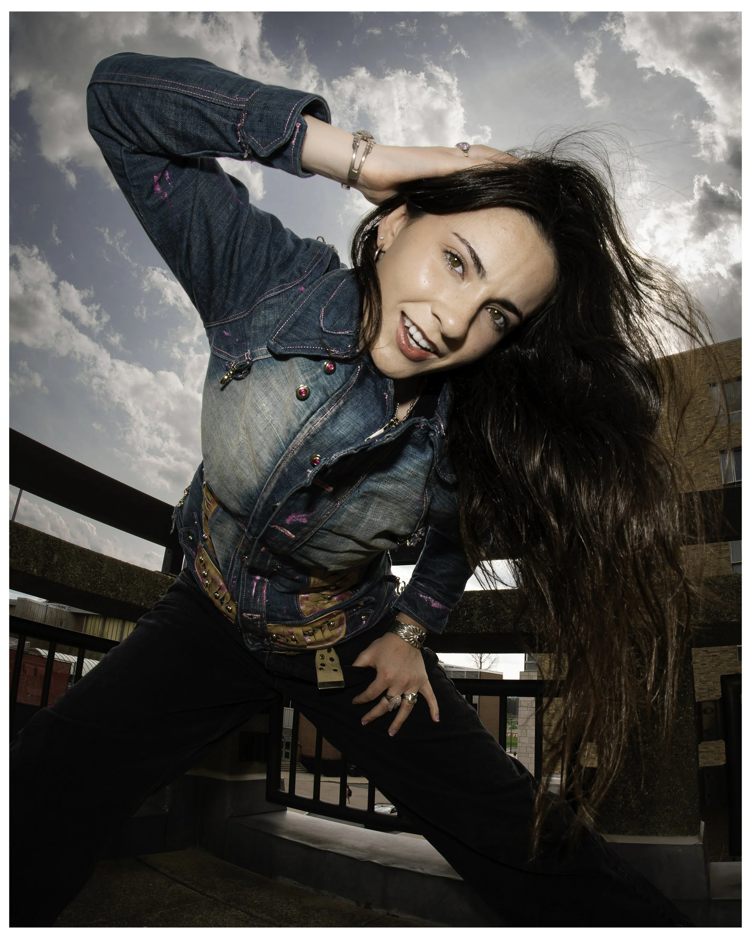 A woman with long dark hair and light skin posing outdoors against a cloudy sky, wearing a denim jacket, black pants, and jewelry.