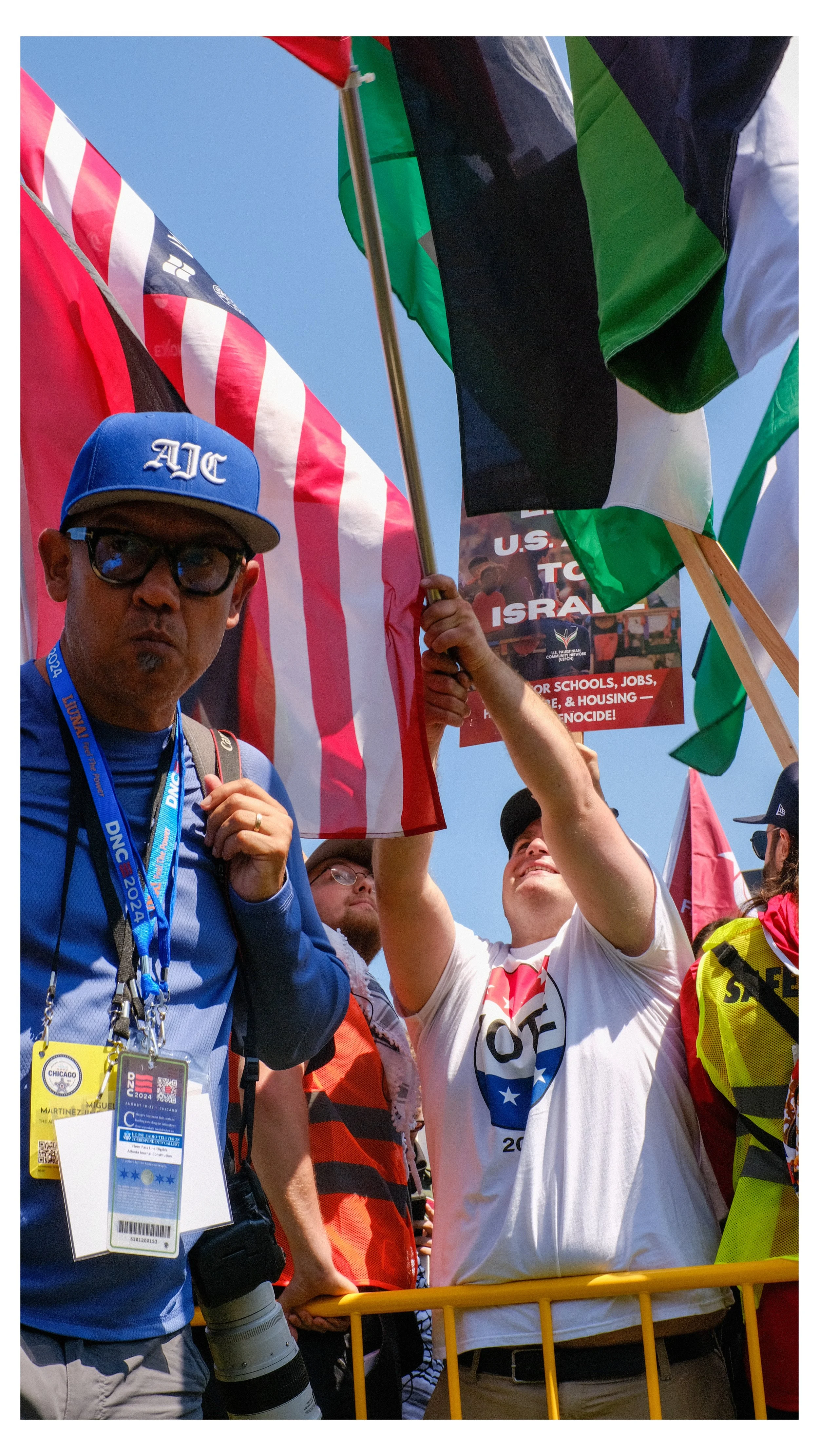 A group of people at a rally or protest, holding flags and signs. One person is wearing glasses, a blue cap with 'AJC' on it, and has a camera with a long telephoto lens. Others are holding flags and a sign that mentions U.S. and Israel, with message